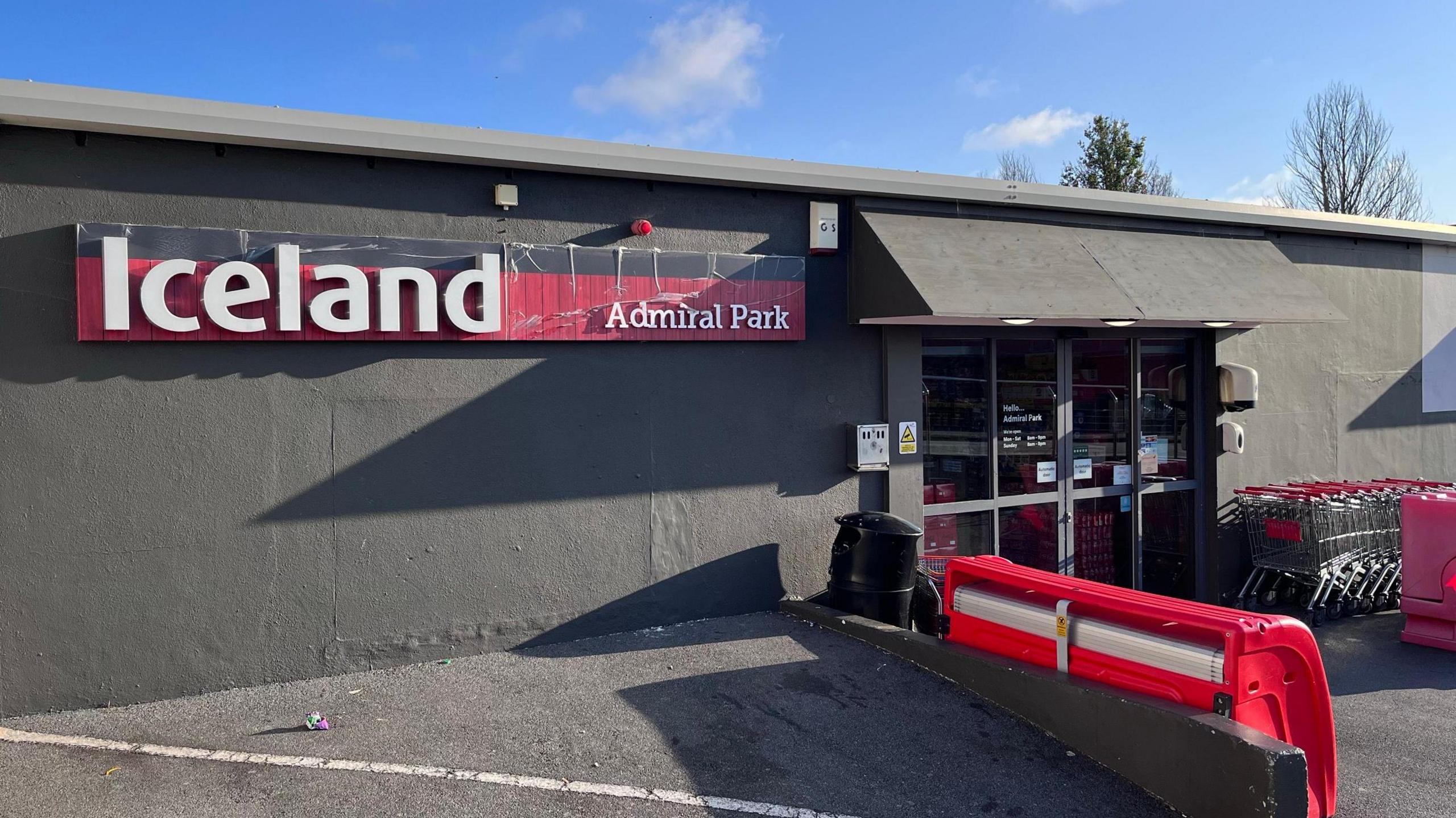 A small Iceland supermarket, with trollies outside. The building is painted dark grey as are the frames of the doors and there is a red Iceland sign on the left of the building also bearing the words 'Admiral Park'.