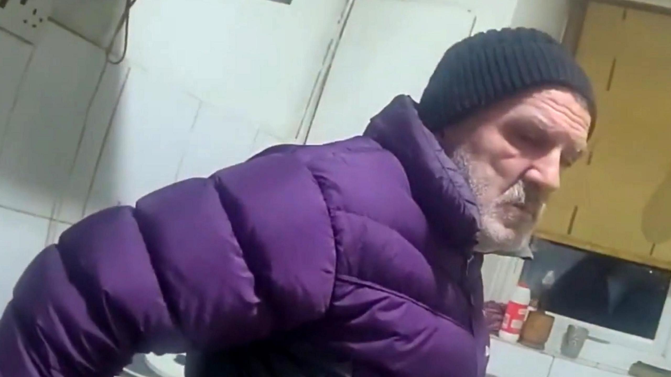 Still from police bodycam footage of Eugeniu Neamtu wearing a purple puffer jacket and black beanie hat. He has a grey beard and is standing in a room