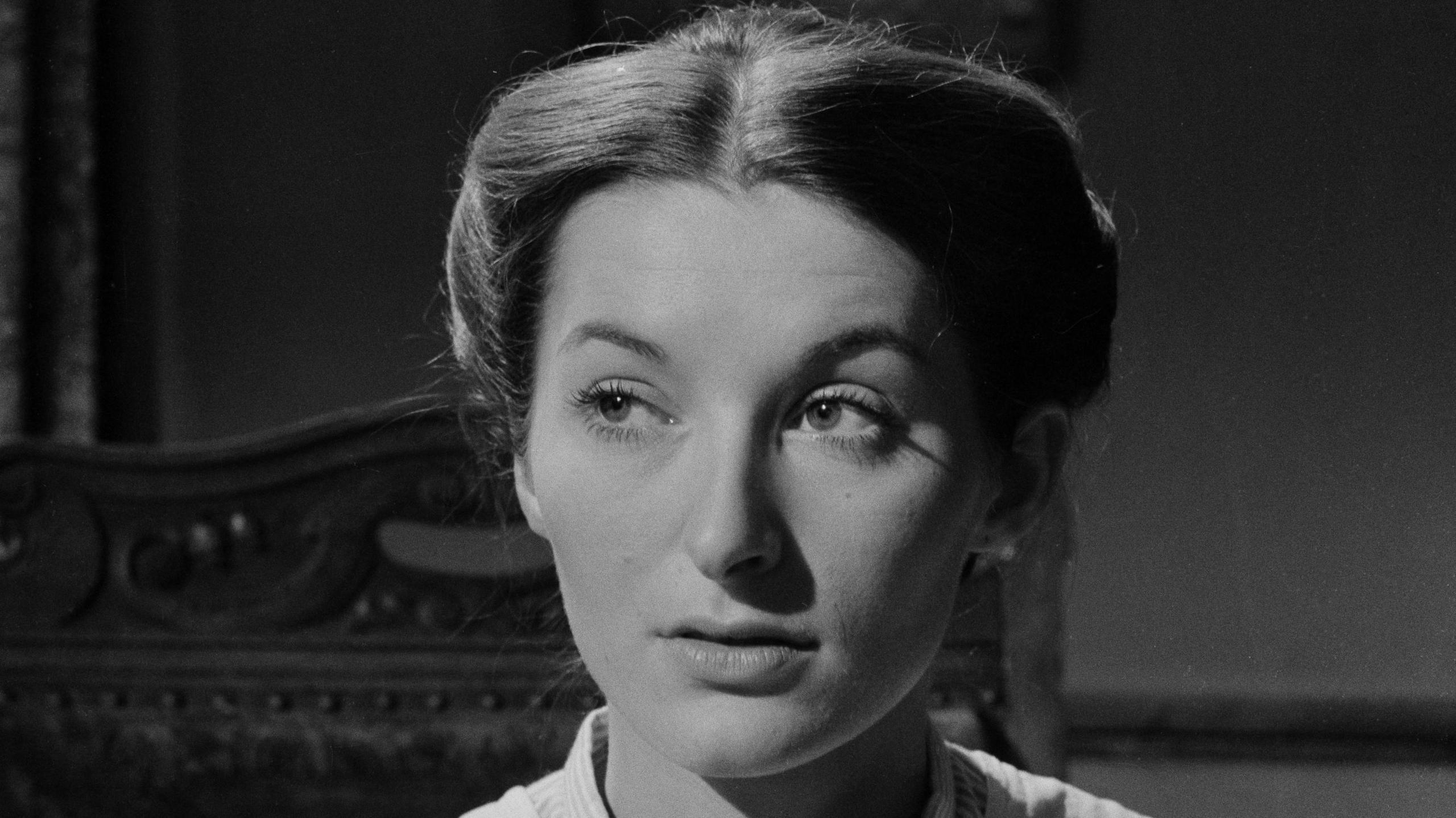 The actress Vivien Heilbron in close-up black and white as Chris Guthrie, the main character in Sunset Song. Her hair is in a tight bun and she is sitting in a wooden chair looking to the left. She looks quizzical.