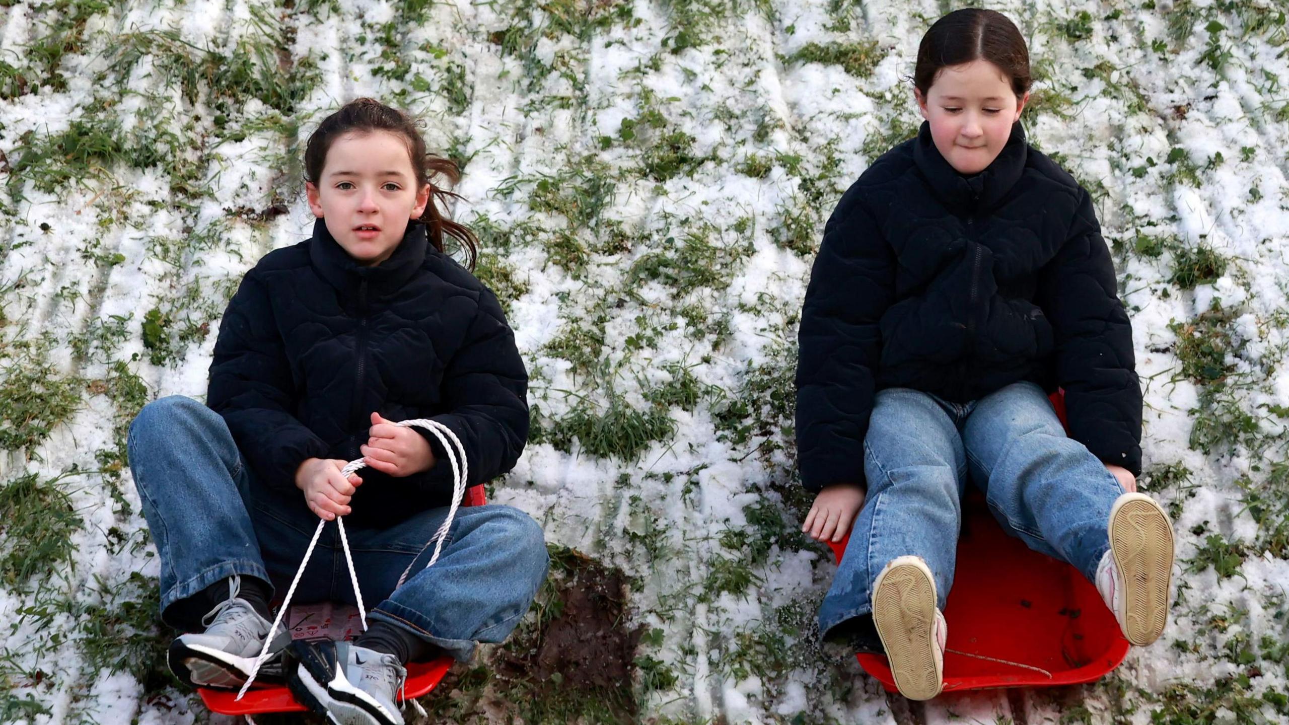 Two girls in red sleighs riding down a snow covered hill. They are both wearing jeans and black hoodies. Snow is sitting on the grass.