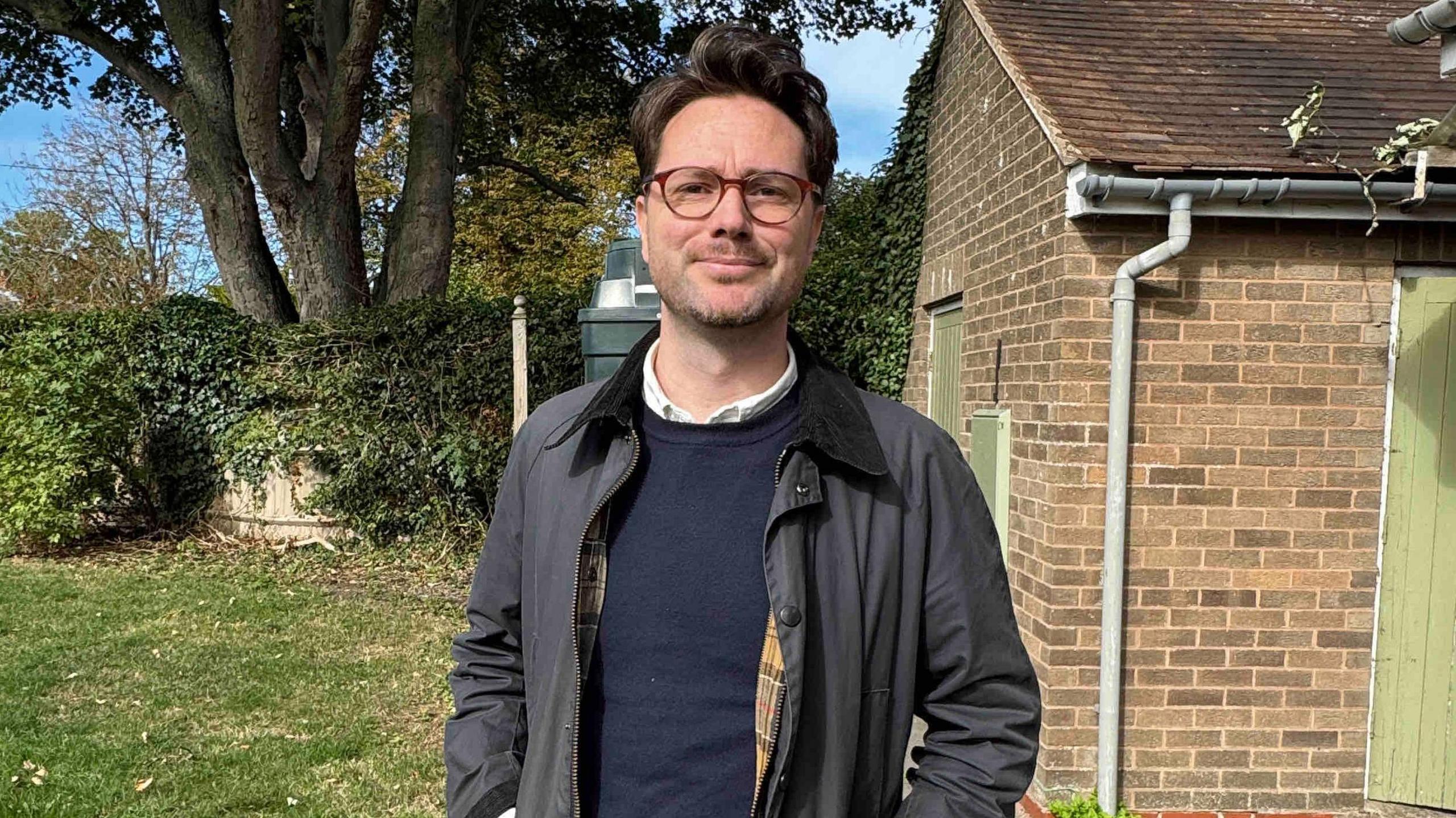 A man with short black hair, glasses, a dark blue top and navy blue jacket, stands in a back garden, next to a building with green doors, with his hands in his pockets. Behind him is a fence covered in greenery and behind that is a large tree with four trunks.