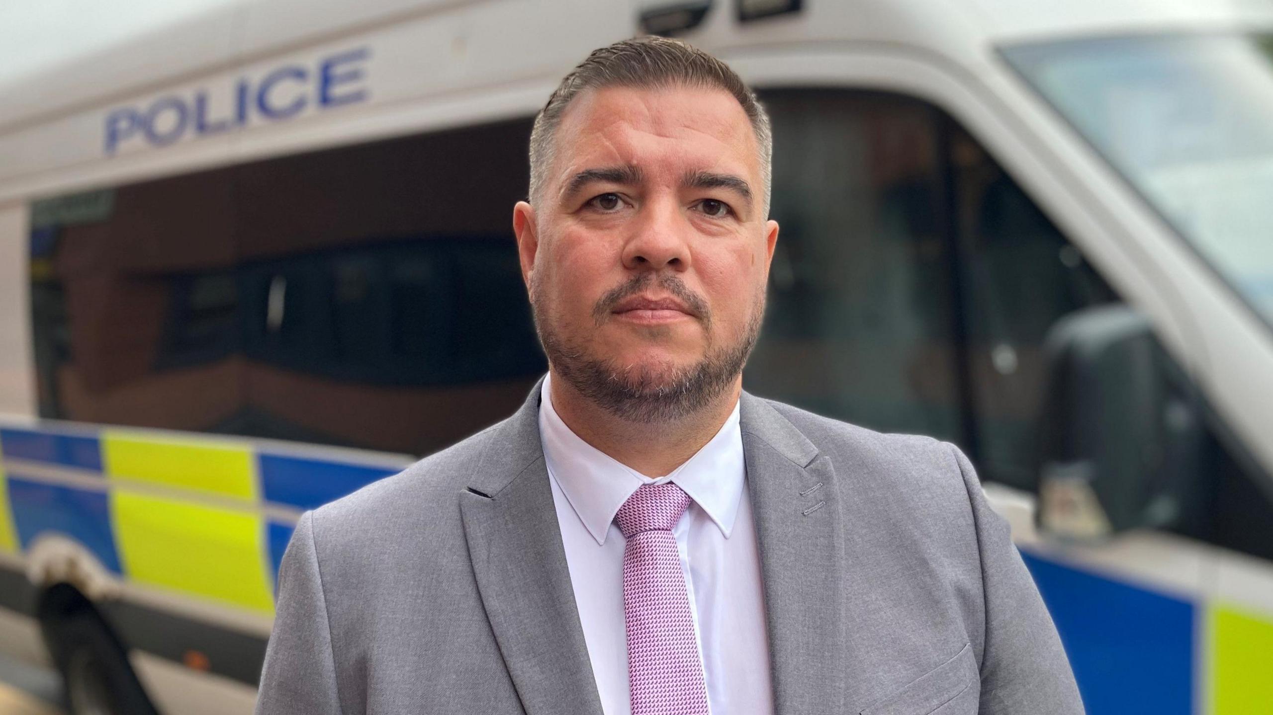Det Ch Insp Stuart Hodgson wears a grey blazer, white shirt and pink tie, with short dark/greying hair and a stubble moustache and beard, and looks into the camera. A police van is parked behind him.