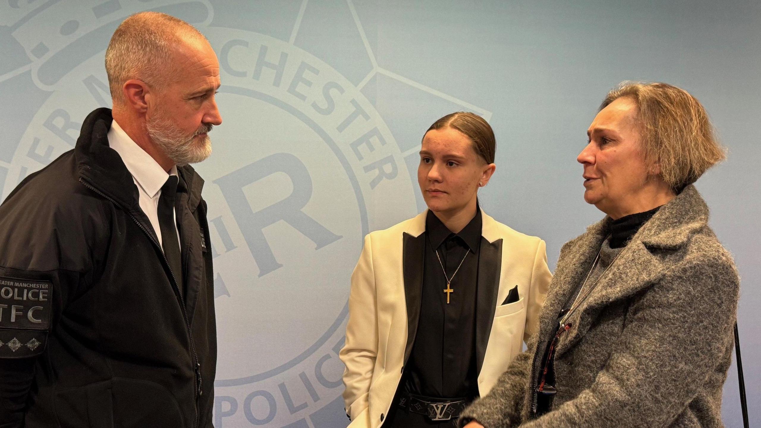 Ch Insp Michael Parker (left) in his Greater Manchester Police (GMP) uniform talking to Wiktoria with brown hair tied back wearing a cream blazer and black top and her mother Claudia with brown bobbed hair wearing a brown coat and grey jumper. They are all side on standing in front of a GMP logo on the wall behind them.
