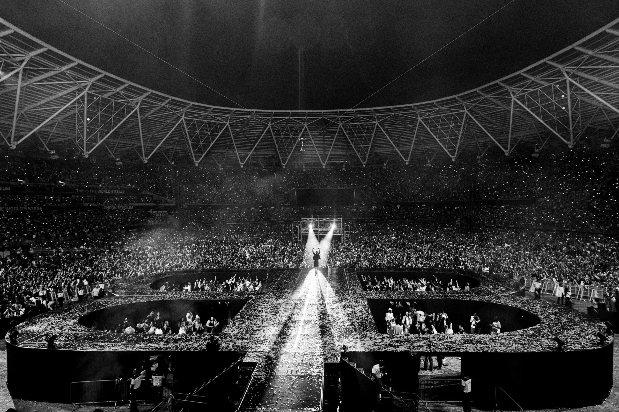 A black and white photo showing Burna Boy on stage in the distance, in a sold out arena with phone lights shining from the crowd.