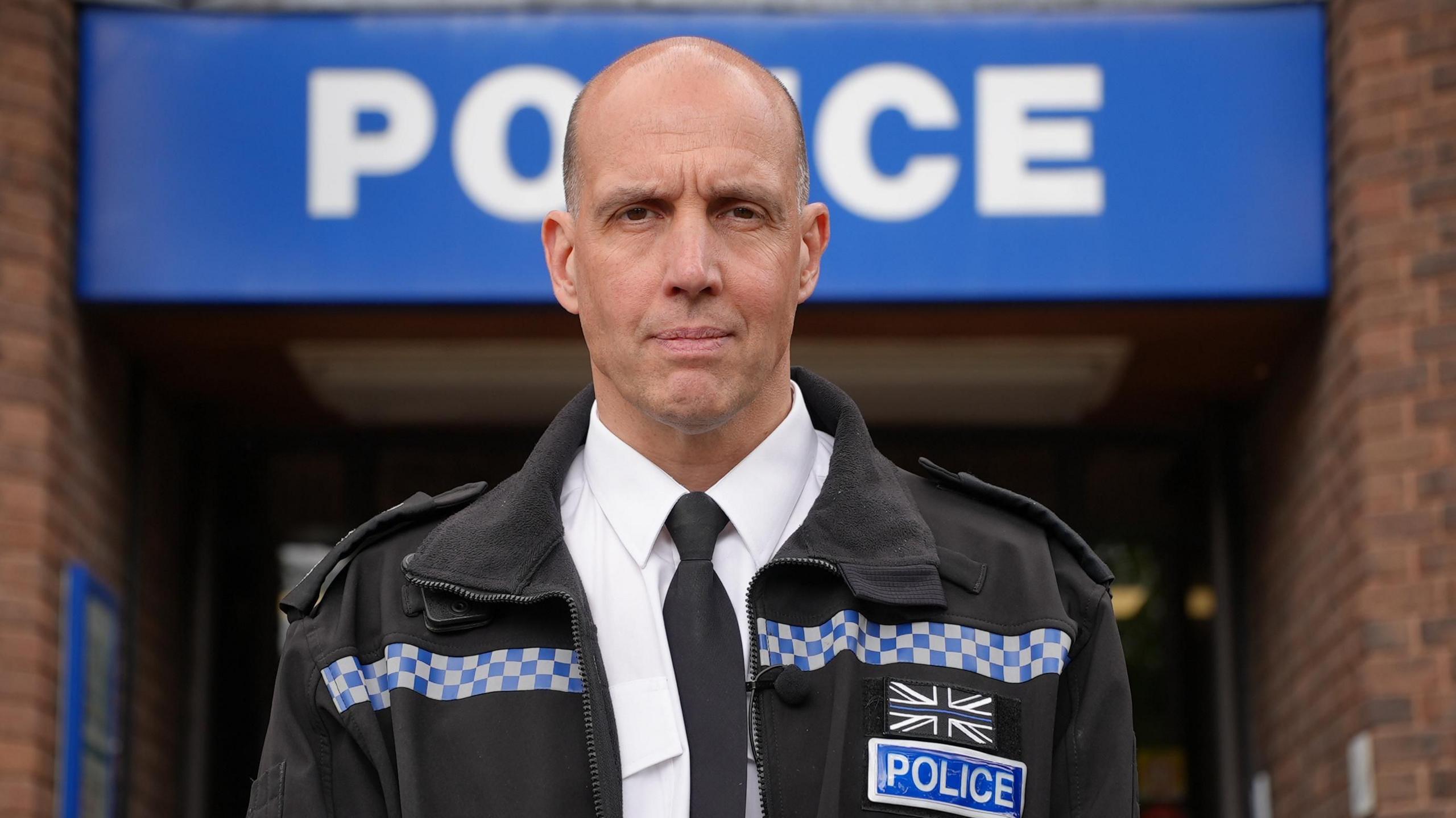 A man with a bald head standing outside a brick building with a large blue sign with 'POLICE' on it in white writing. He's wearing a black jacket with a checked blue and white strip across the chest, and a label reading 'POLICE'.