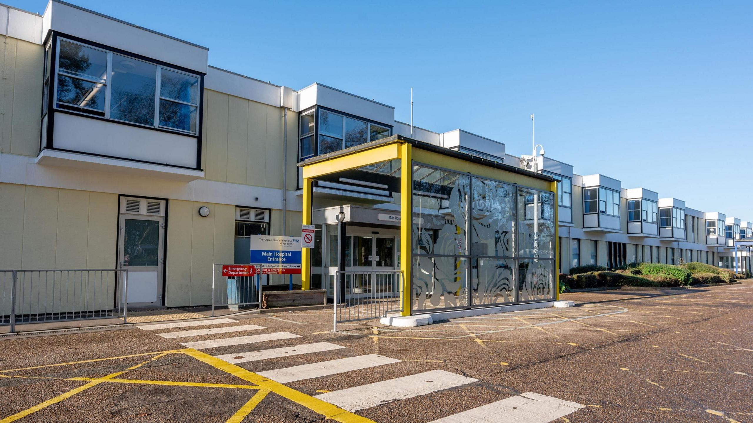 Exterior of Queen Elizabeth Hospital showing a two storey, low-lying building with first floor windos jutting out and a yellow framed and covered entry to the double doors