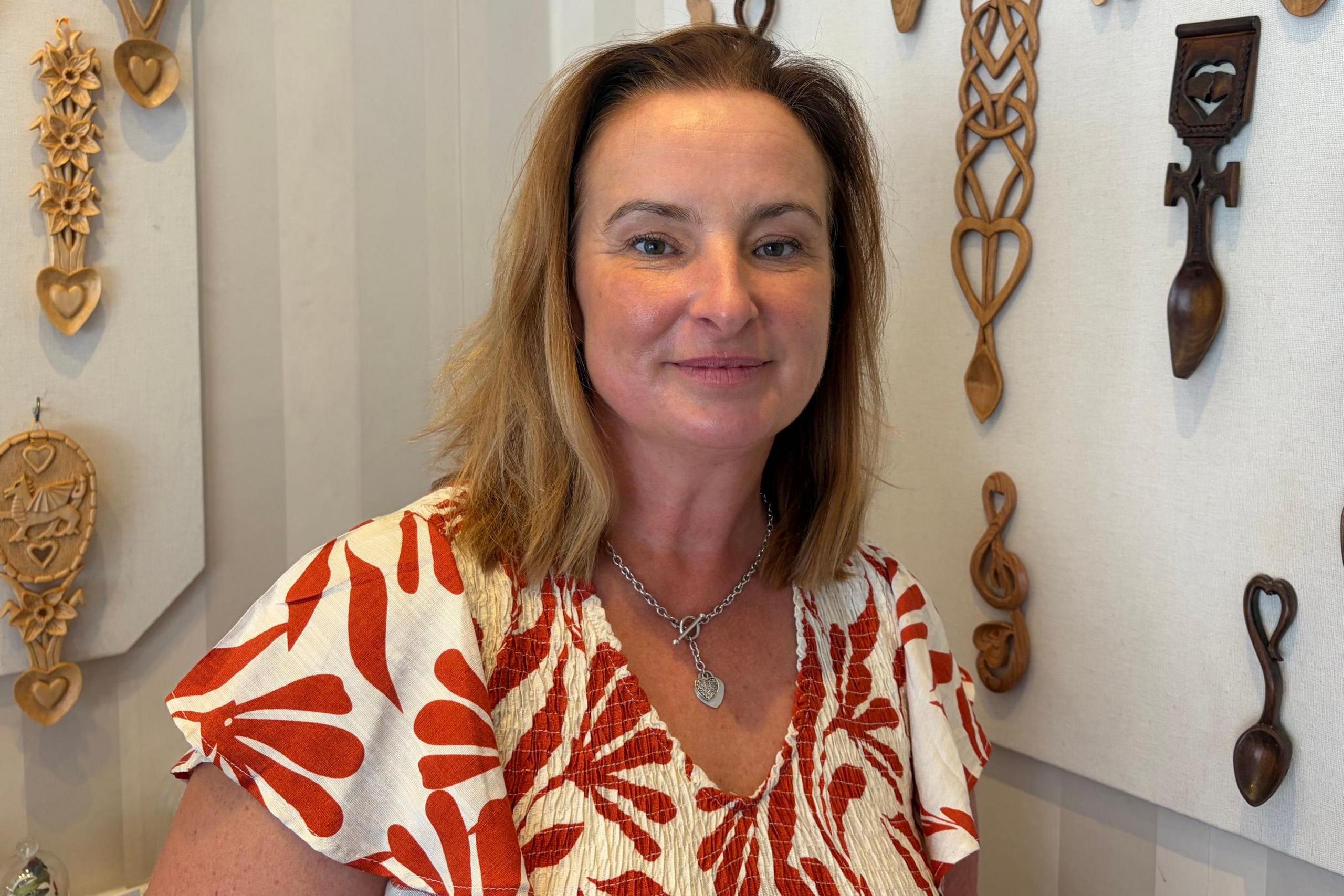Jane, who has light brown hear and wears a floral white and red top, with love spoons attached to a wall in the background.