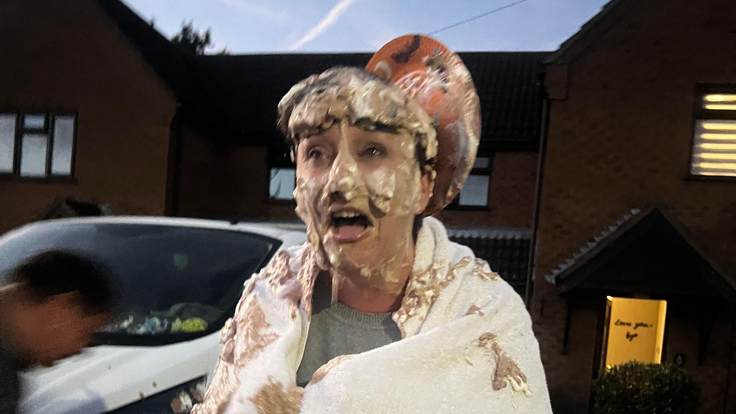 A lady with a grey round collared shirt under a white towel, which is wrapped around her shoulders. The towel is splattered in brown and white squirty cream. An empty paper plate can be seen on the top of the ladies head. She looks fairly shocked. A white car is behind her, and beyond that is a house.