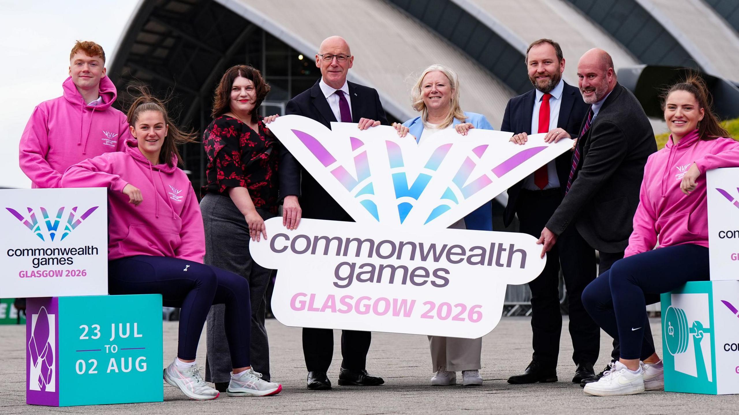 John swinney with a group of officials and athletes holding up a large glasgow commonwealth games sign.