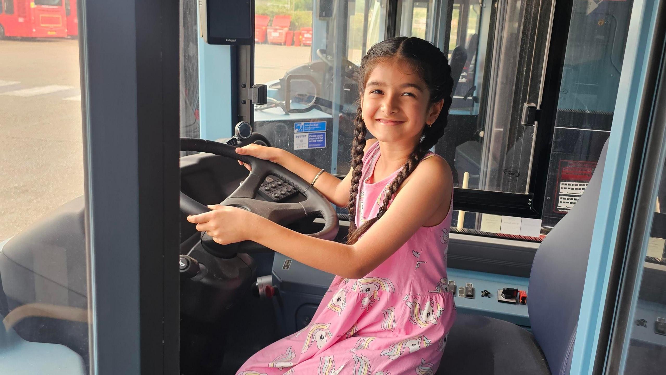 A young girl sits in the drivers seat of a bus in a pink dress 