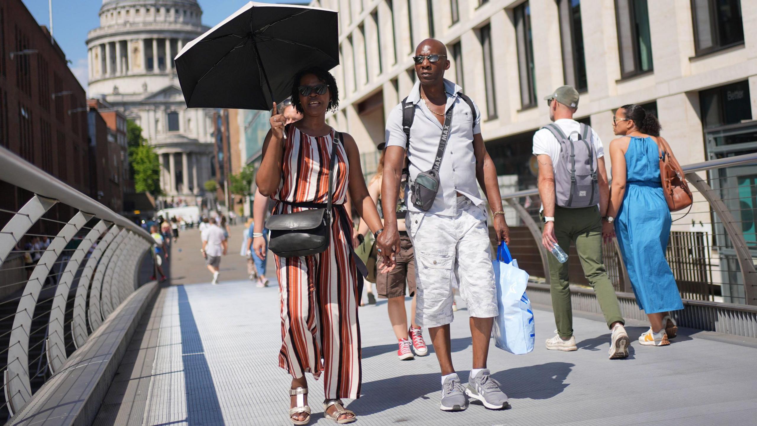 A woman wearing a black, white, yellow, and orange striped jumpsuit holds an umbrella in the sun. A man wearing a grey shirt and white shorts holds her hand and walks with her.