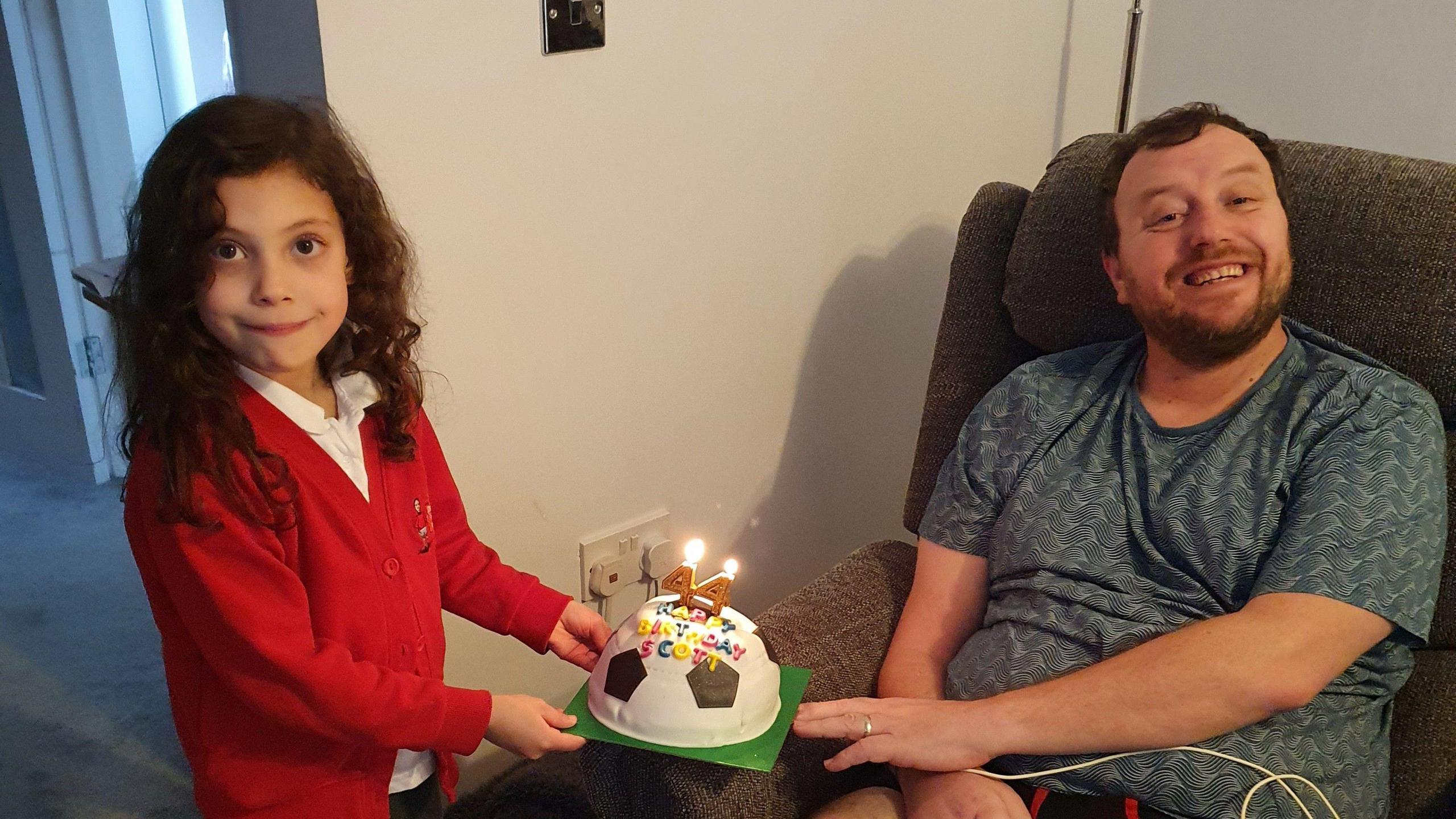 A young girl in a red jumper presents a man sitting in a chair with a football birthday cake with candles in the shape of a 44.