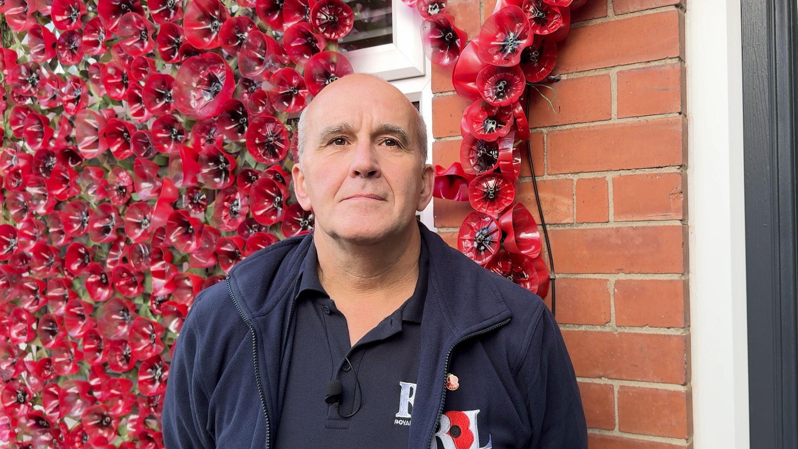 A bald gentleman staring at the camera, with a poppy display behind him. He's wearing a navy Royal British Legion Jacket and is wearing a pinned poppy. 