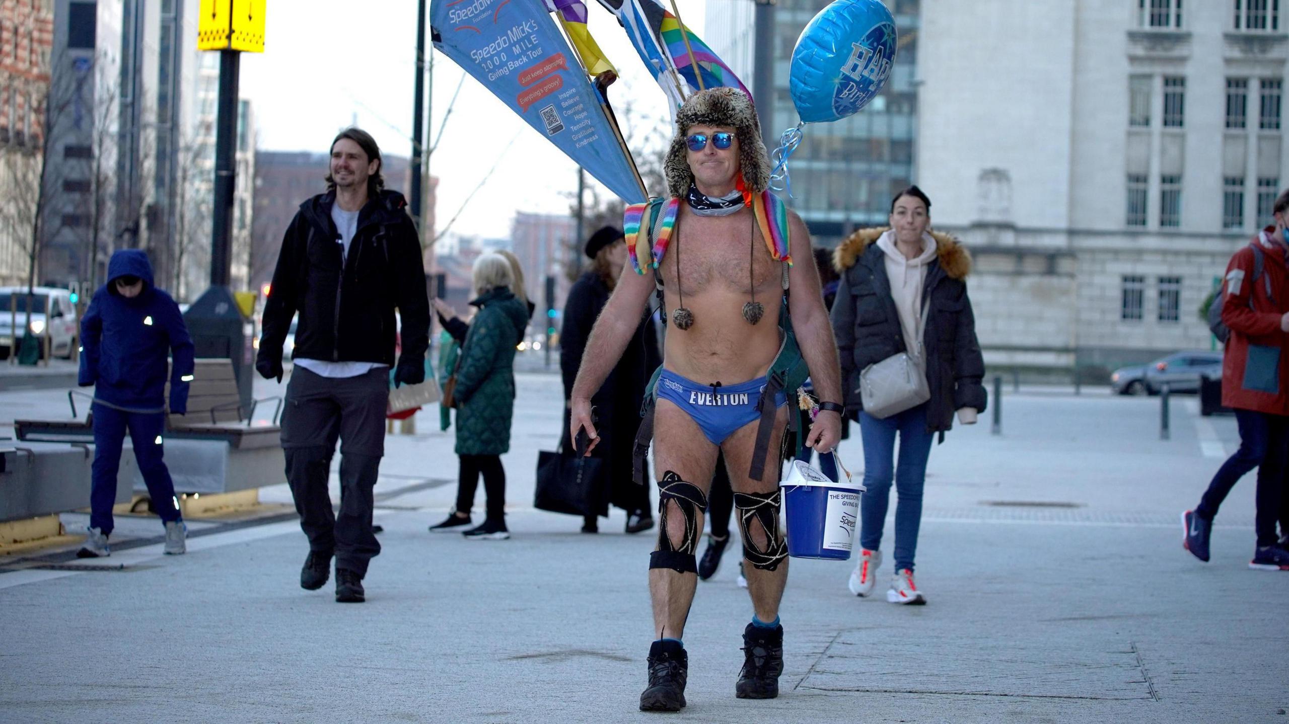 Speedo Mick, wearing a brown furry hat, a rucksack and blue swimming trunks, walks through the streets in Liverpool with a bucket to collect donations