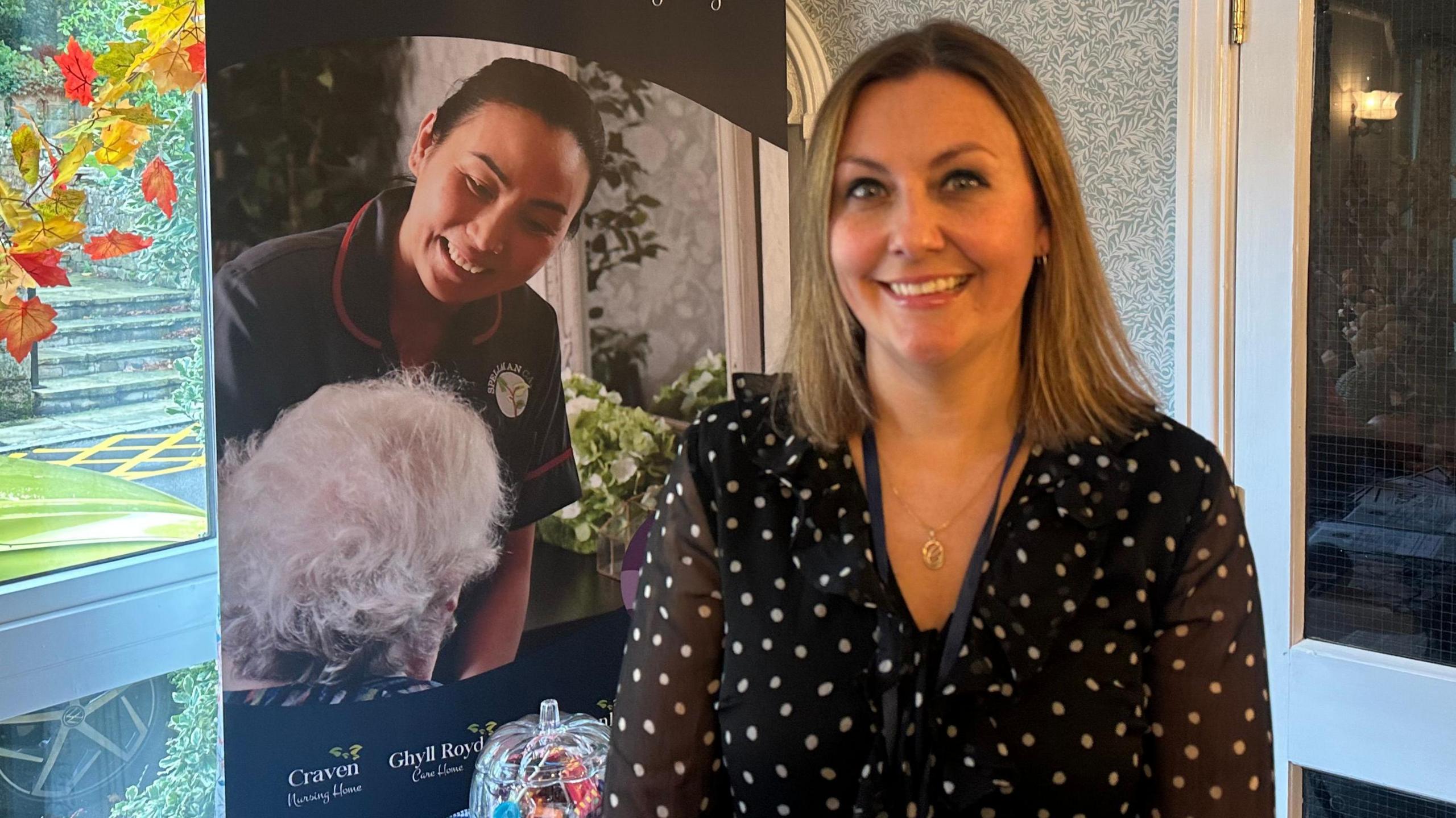 A woman in a black top smiles. Behind her is a banner that reads "Spellman care". A window is next to her which show autumn leaves outside.