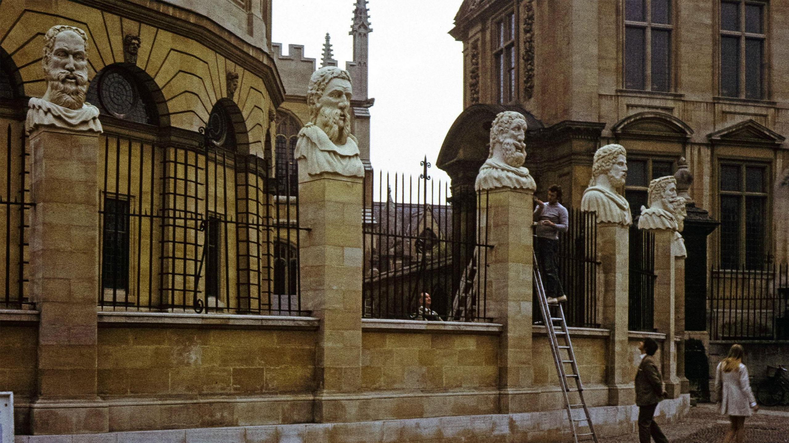 Michael Black is pictured sculpting the head figurines known as the Emperor's Heads mounted on pillars outside the Sheldonian Theatre in Oxford. A man can be seen at the foot of the ladder watching the sculptor at work.