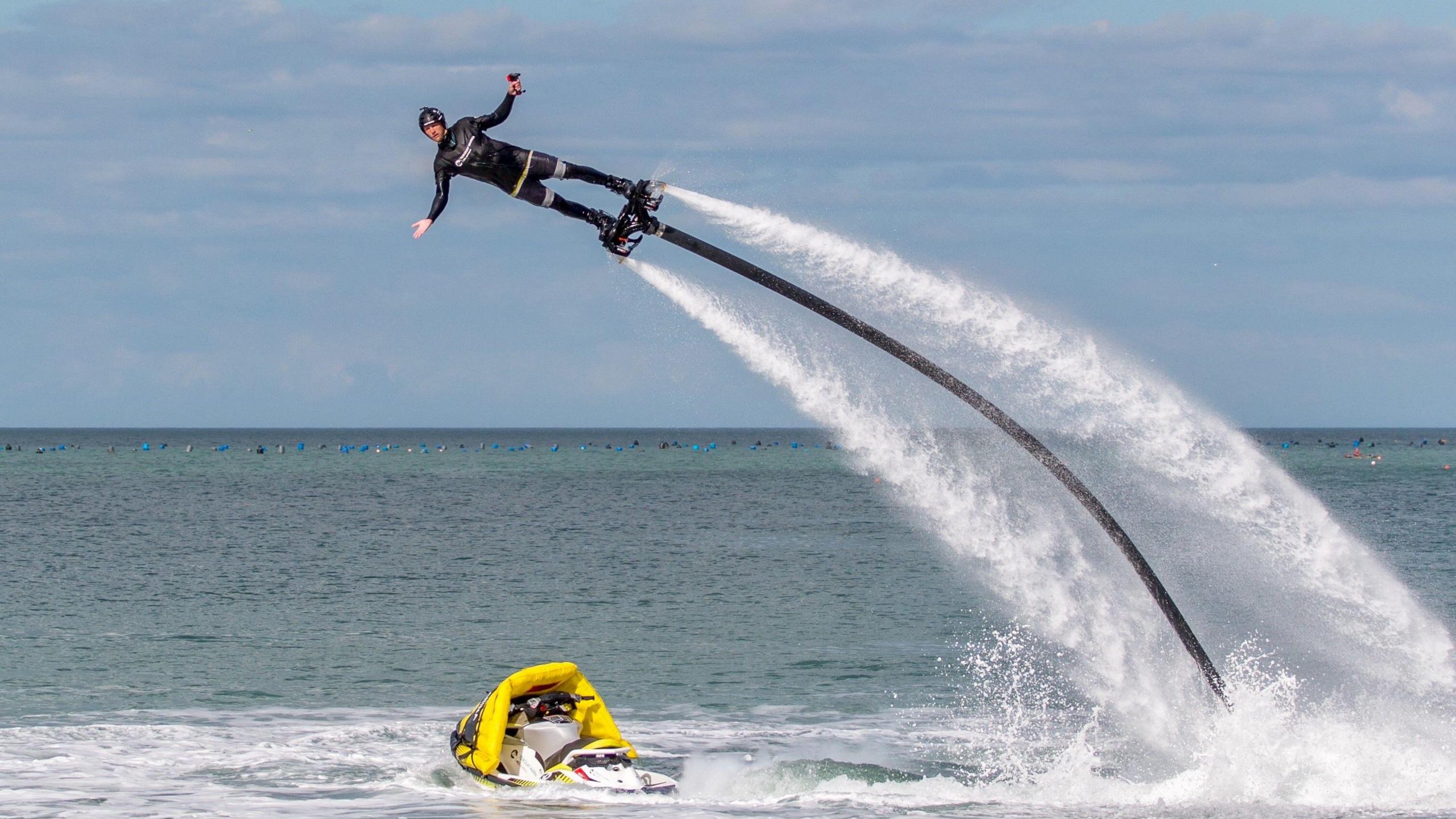 A man in a black wetsuit is pictured on a flyboard with water shooting out the bottom.