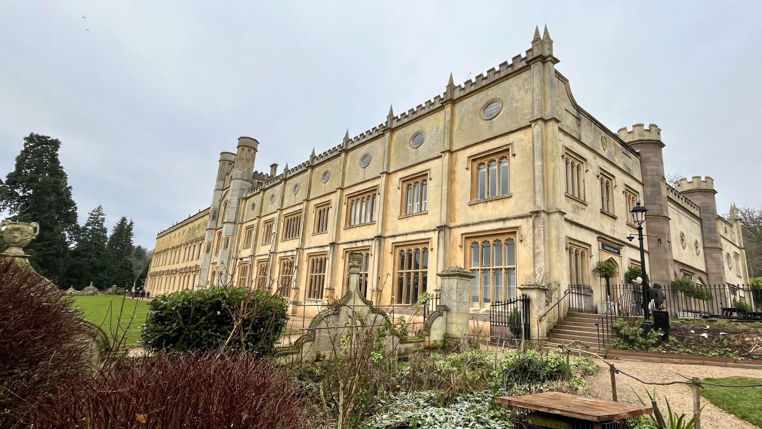 A wide shot of Ashton Court Mansion taken from just below the building. In the foreground several different shrubs are visible. The exterior of the building is a faded yellow and there is a section of green lawn visible in front of it. At the side a set of steps lead up to a courtyard