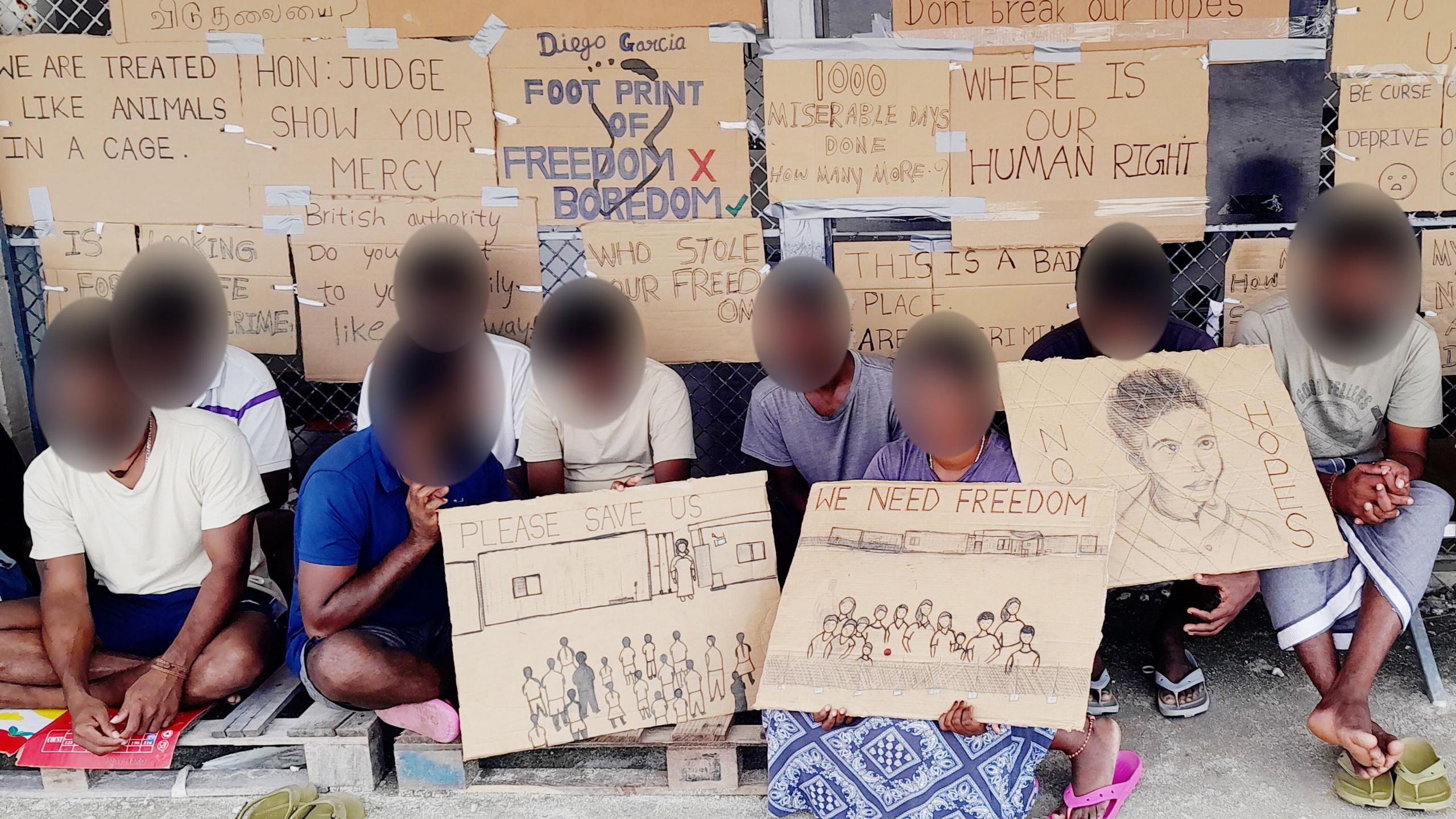 Tamil migrants on Diego Garcia, with their faces blurred, holding protest signs reading "please save us" and "we need freedom", with other signs attached to a fence behind them