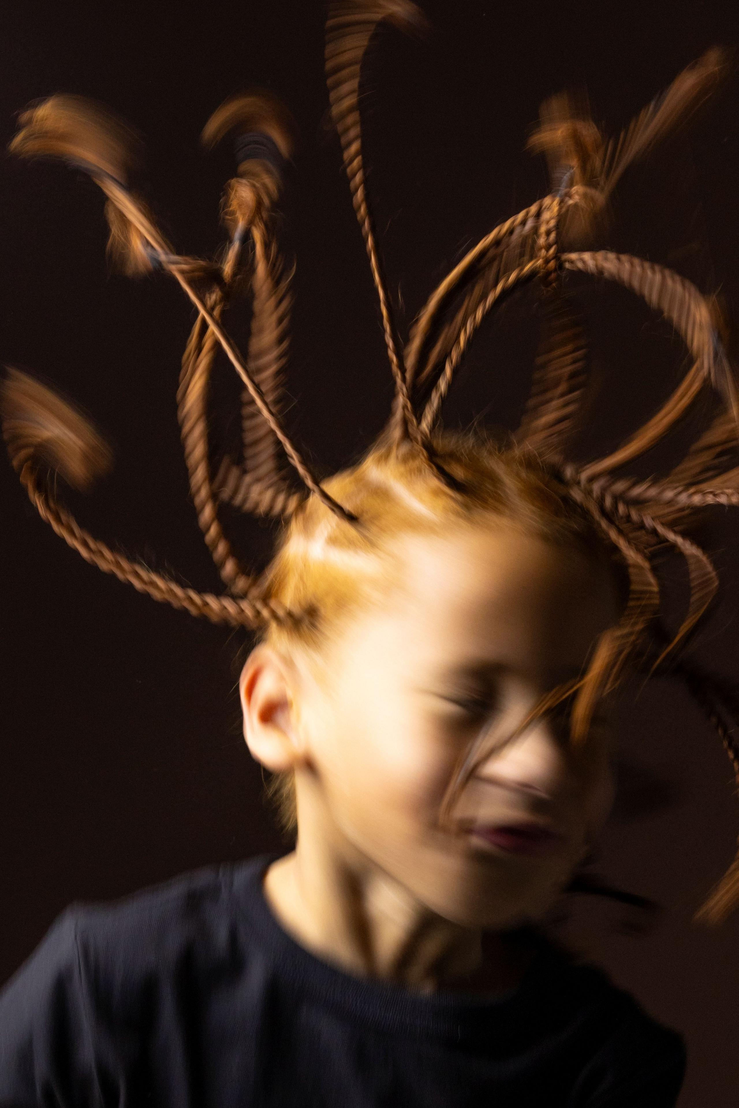 Artistic close-up portrait of a child with multiple braided strands of hair extended outward in motion, creating a dynamic blurred effect. The child wears a dark top and is photographed against a dark brown background with warm studio lighting that highlights the movement and texture of the braids.