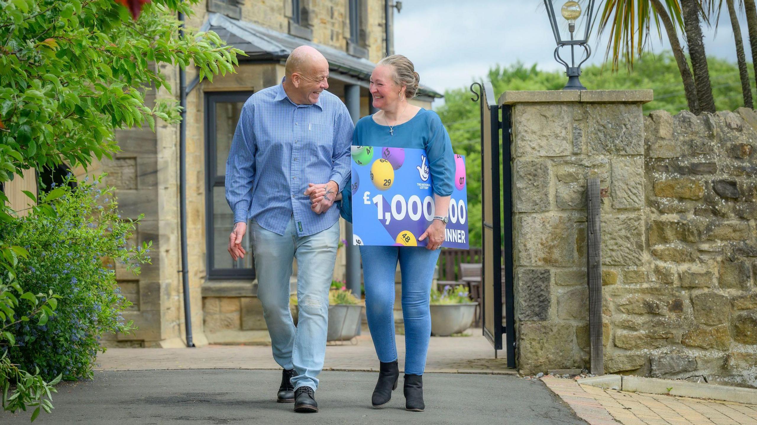 Mr and Mrs Gillings smile as they walk hand in hand. Mrs Gillings is holding a sign that reads "£1,000,000 winner" under her left arm.
