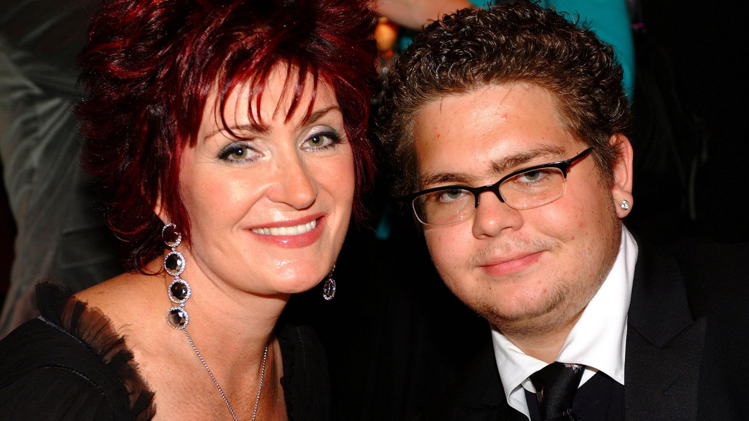 A close-up of Sharon Osbourne, left, with a younger Jack, right. Sharon is wearing a black low cut top and large dangling black earrings. Her hair is red and cut short and she is smiling. Jack is wearing a black suit and tie with white shirt, wire-framed glasses and diamond studs in his ears.