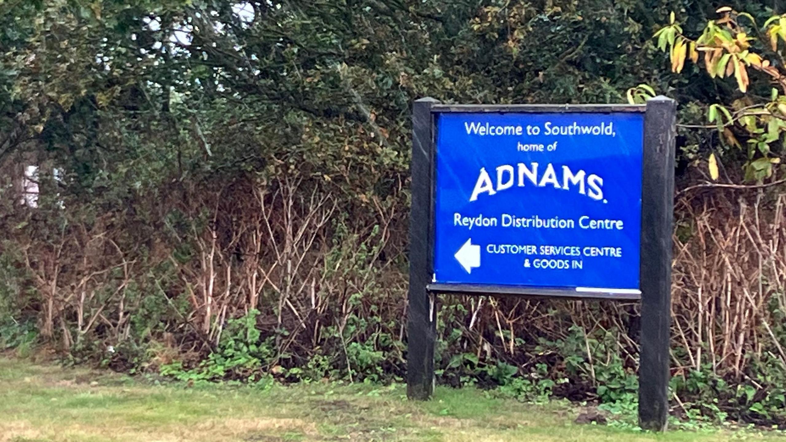 Blue sign reading "Welcome to Southwold, home of Adnams Reydon Distribution Centre" in front of green foliage 