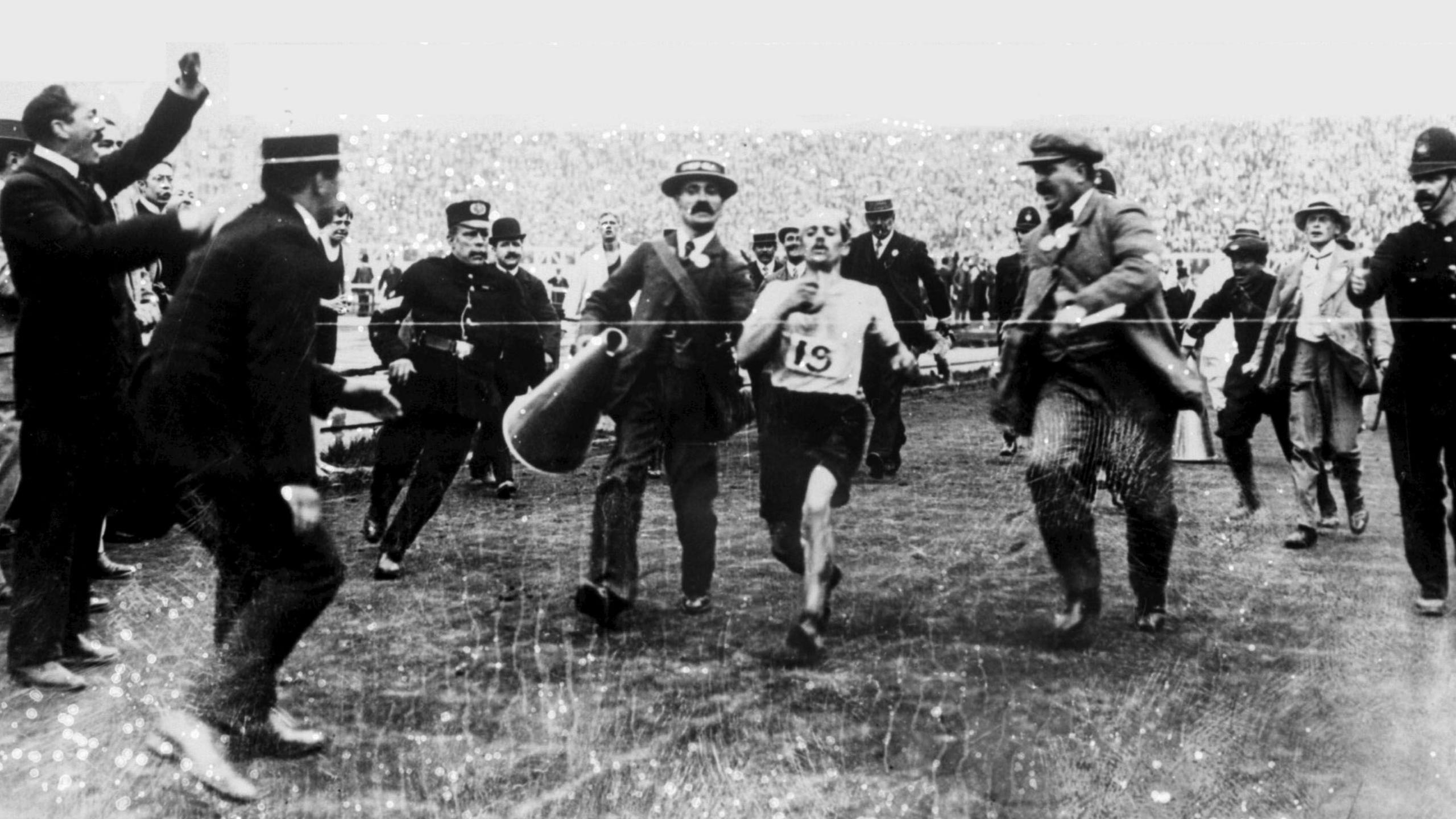 A black and white photo shows Pietri about to break the tape at the finish line of the 1908 marathon. Officials stand to either side, some with their arms in the air and beside him and man in a suit holds a large loud hailer.