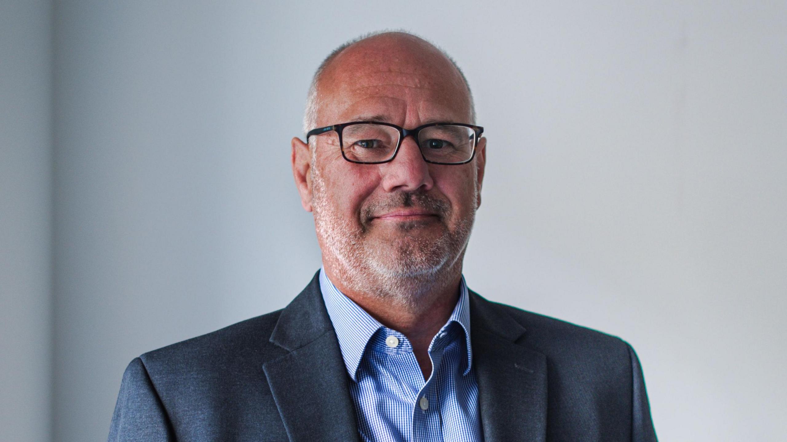 Darryl Goddard. He is wearing a grey suit, blue shirt and glasses. He has short grey hair and stubble. 