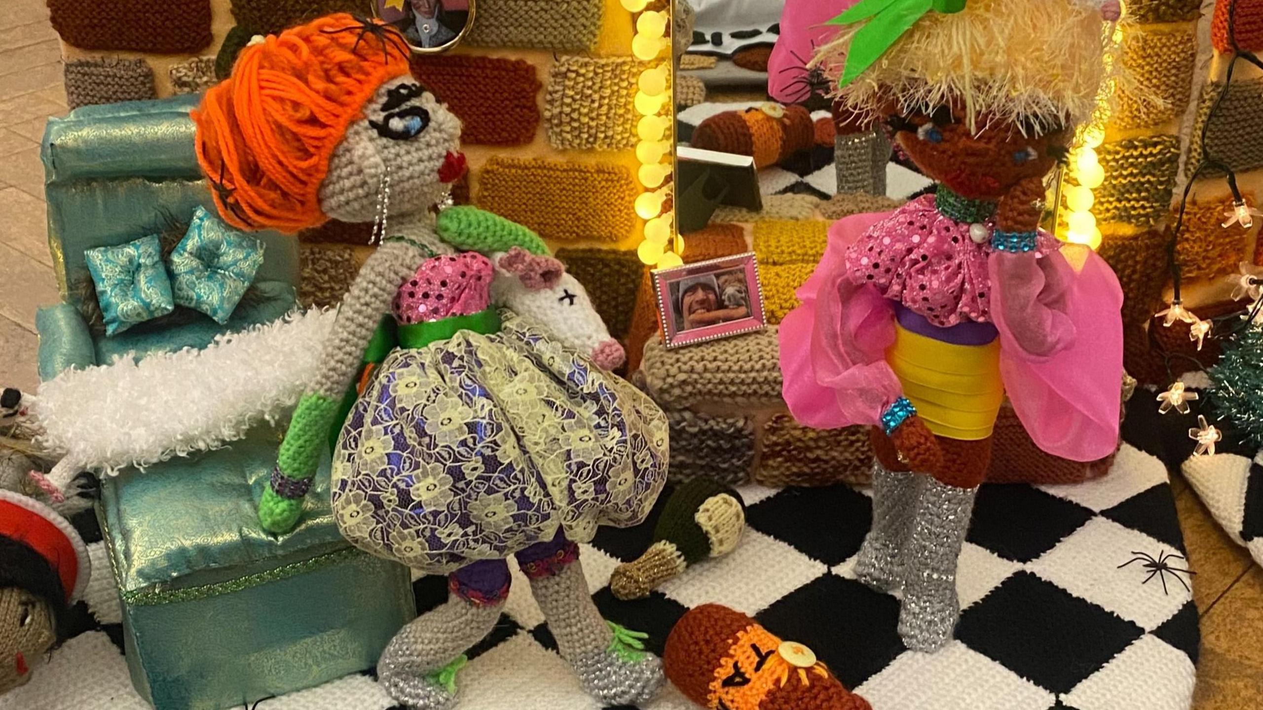 Two Cruel Sisters made out of wool. They are dressed in multi-coloured outfits with colourful hair. They are on a black and white floor that has a fake tan bottle on it. They are beside a mirrored light. 