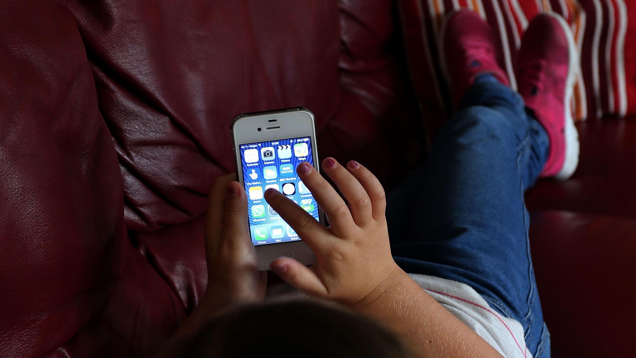 Close up of a child's hand tapping on a mobile phone screen with apps visible.