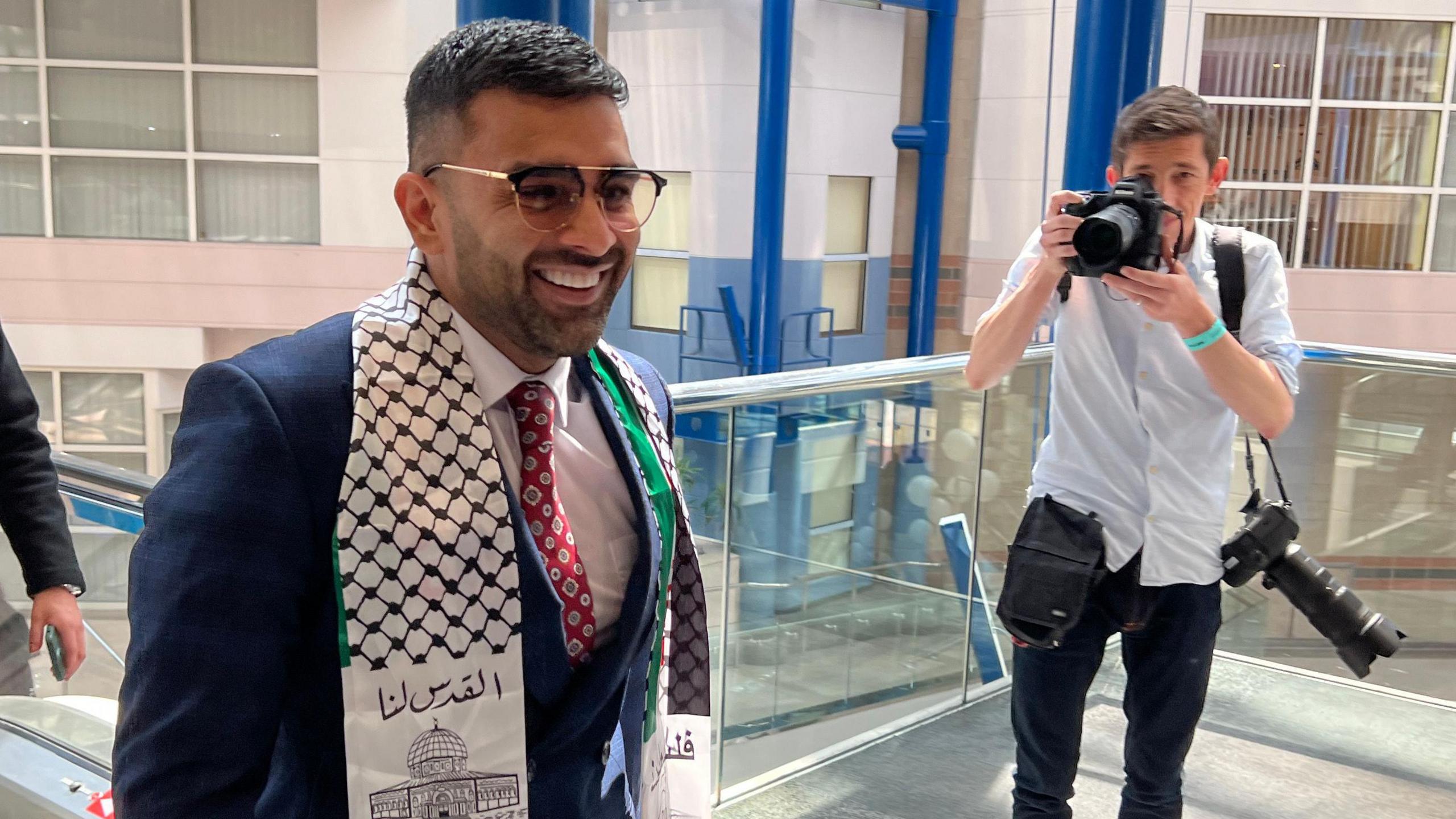 A man wearing a blue suit and white scarf. He is smiling. A photographer in a white shirt and dark trousers stands to one side, taking a picture of him