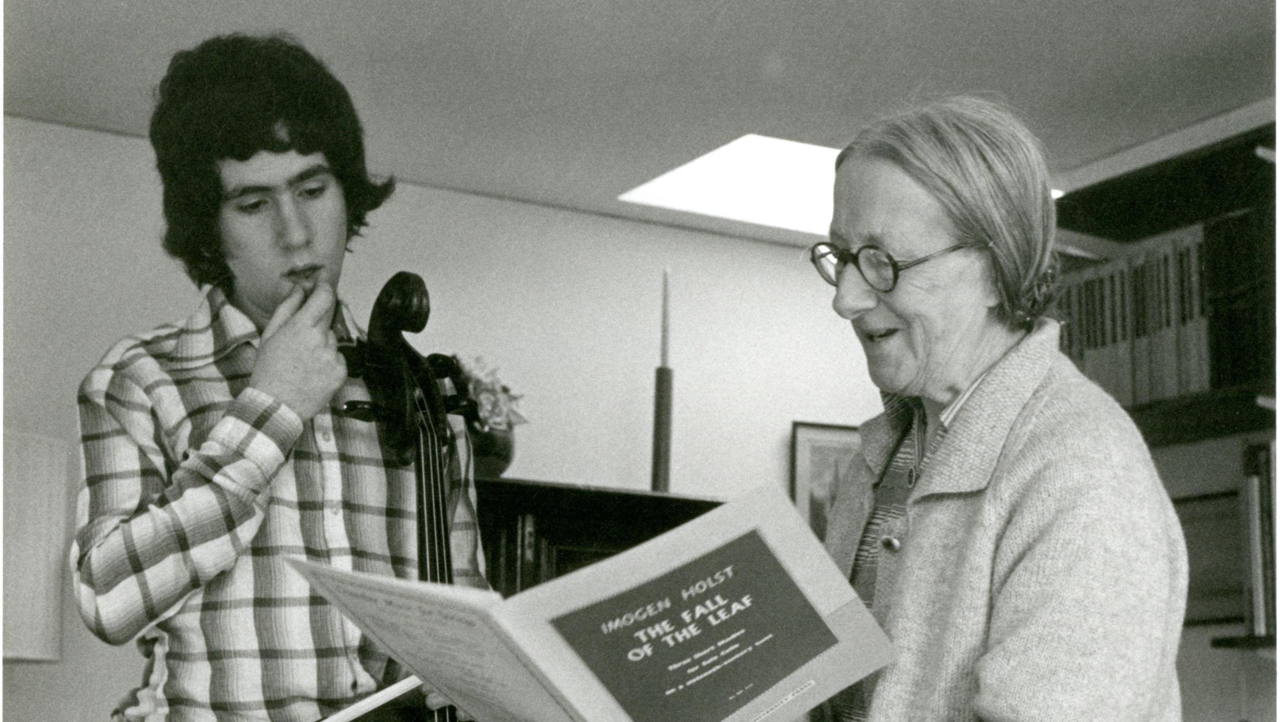 Imogen Holst readers a music sheet as Steven Isserlis stands next to her also looking at the sheet music. Imogen wears glasses and a cardigan while Steven has dark curled hair and wears a chequered shirt with a cello next to him. The image is in black and white.