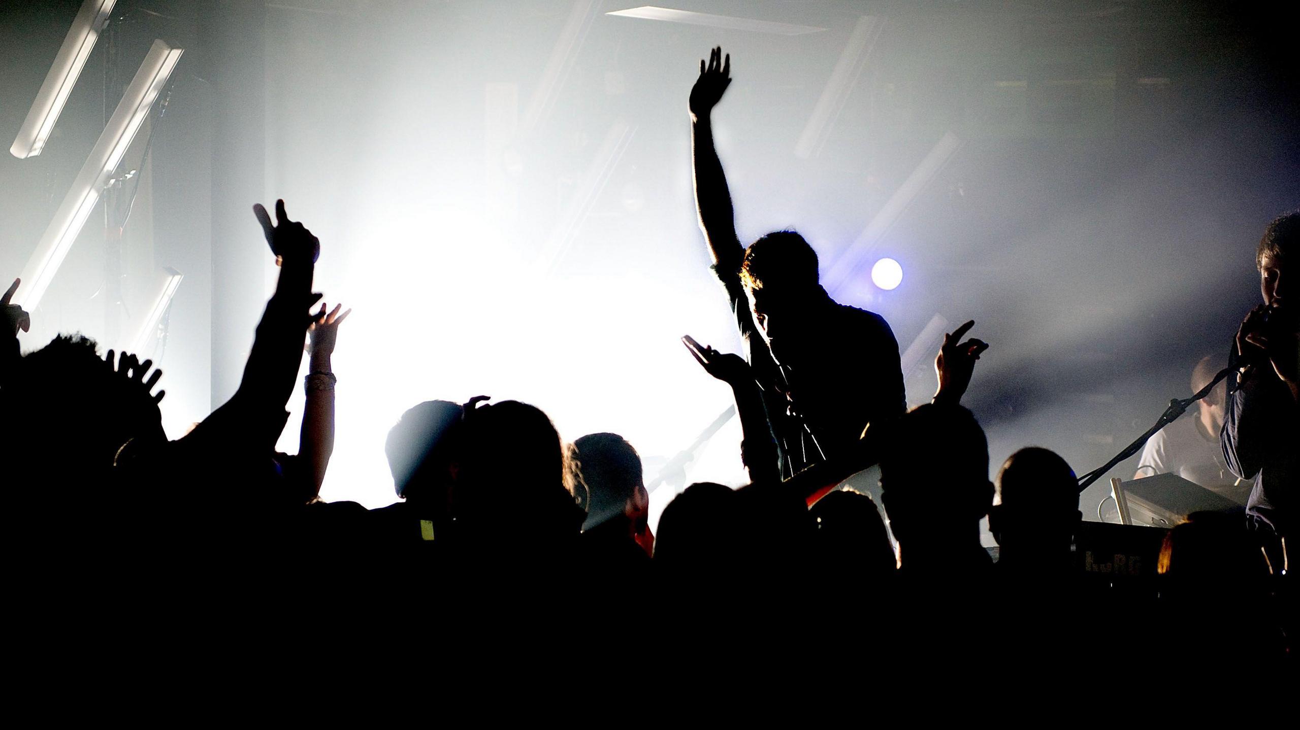 Clubbers holding their arms in the air are pictured in silhouette as they watch a band perform, with a guitarist also in silhouette holding his right arm in the air. The stage is backlit.