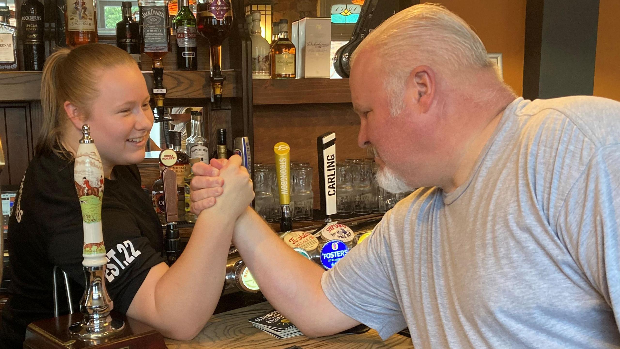 Eve is in a black T-shirt and her dad is leaning over the counter of a bar in a grey t-shirt to arm wrestle her.
