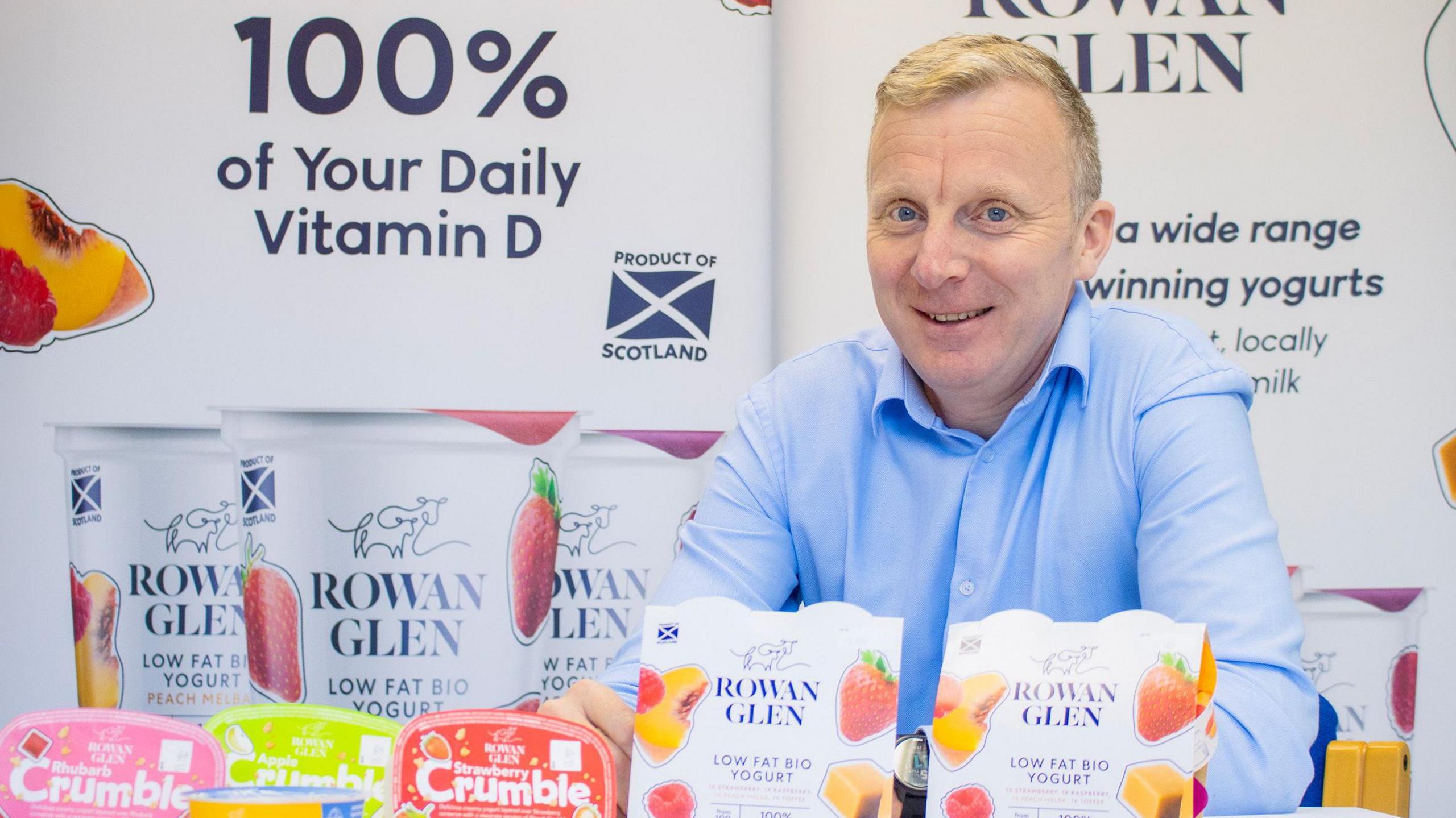 A man in a blue shirt with short blond hair sits behind yoghurt products with the Rowan Glen name on them