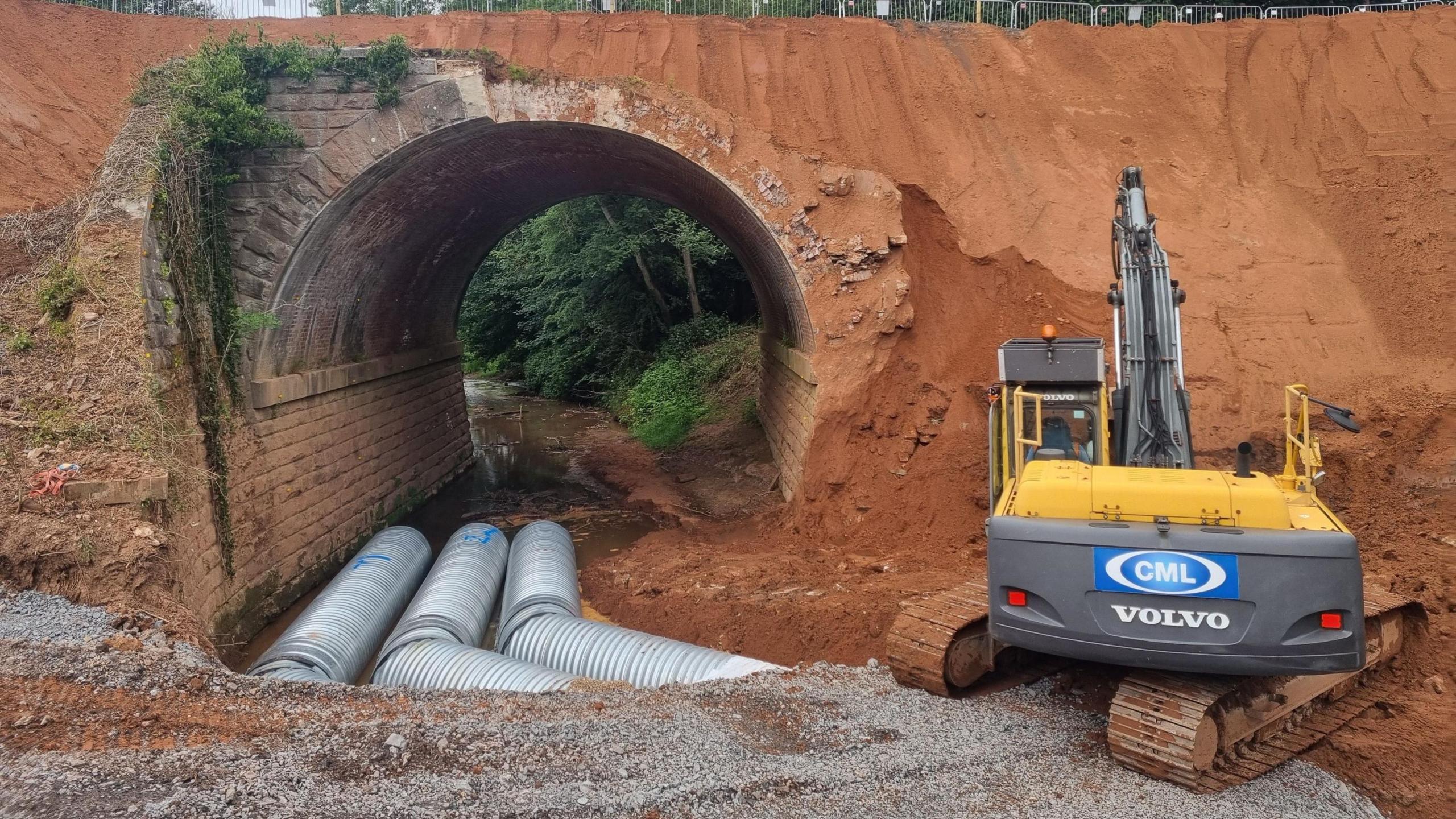 A red earth tunnel with three metal tubes running through it and a yellow and black mechanical digger in the foreground