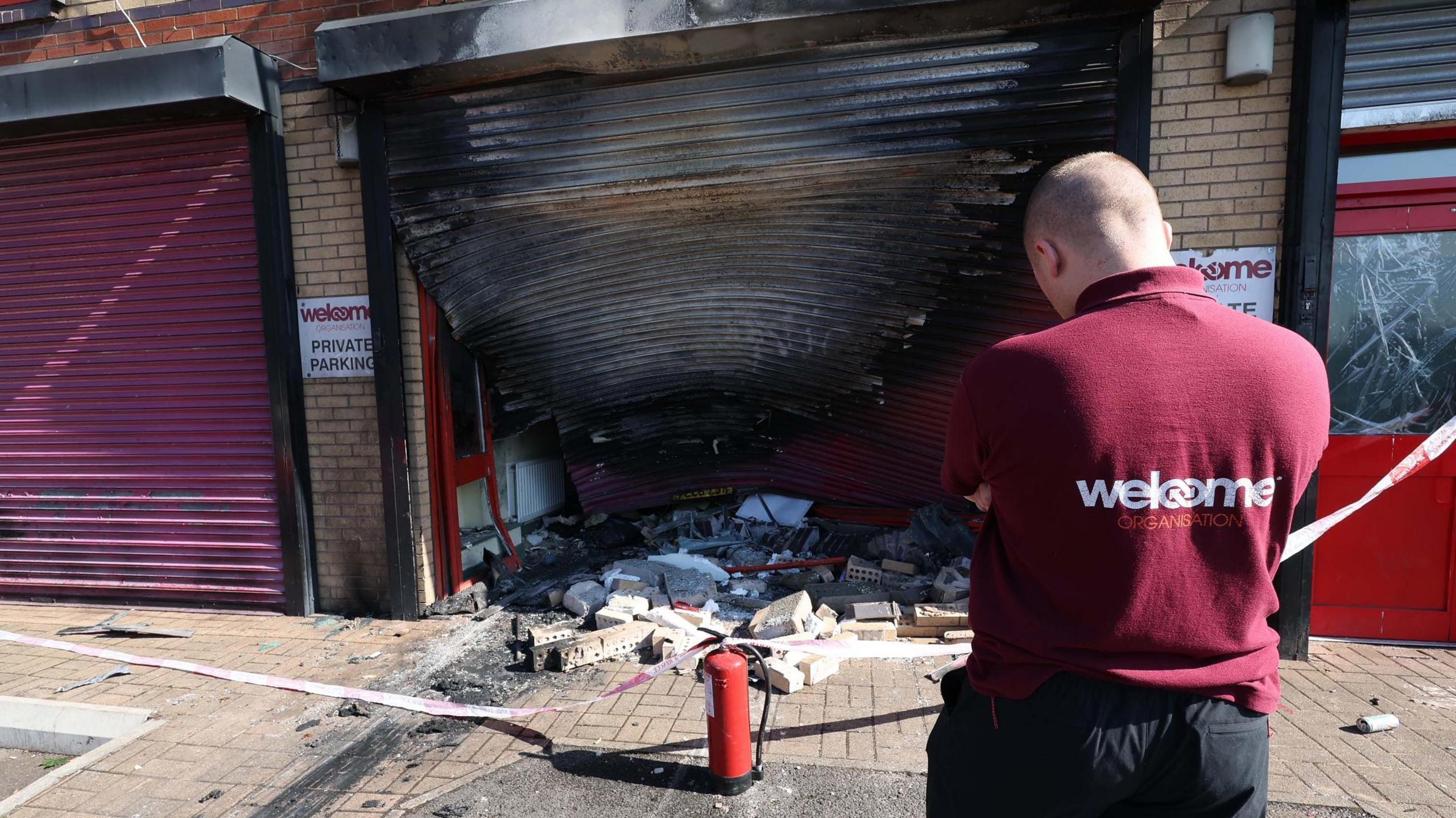 The exterior of the Welcome Organisation building with a burned garage door which is warped as a worker in a maroon polo shirt surveys the damage