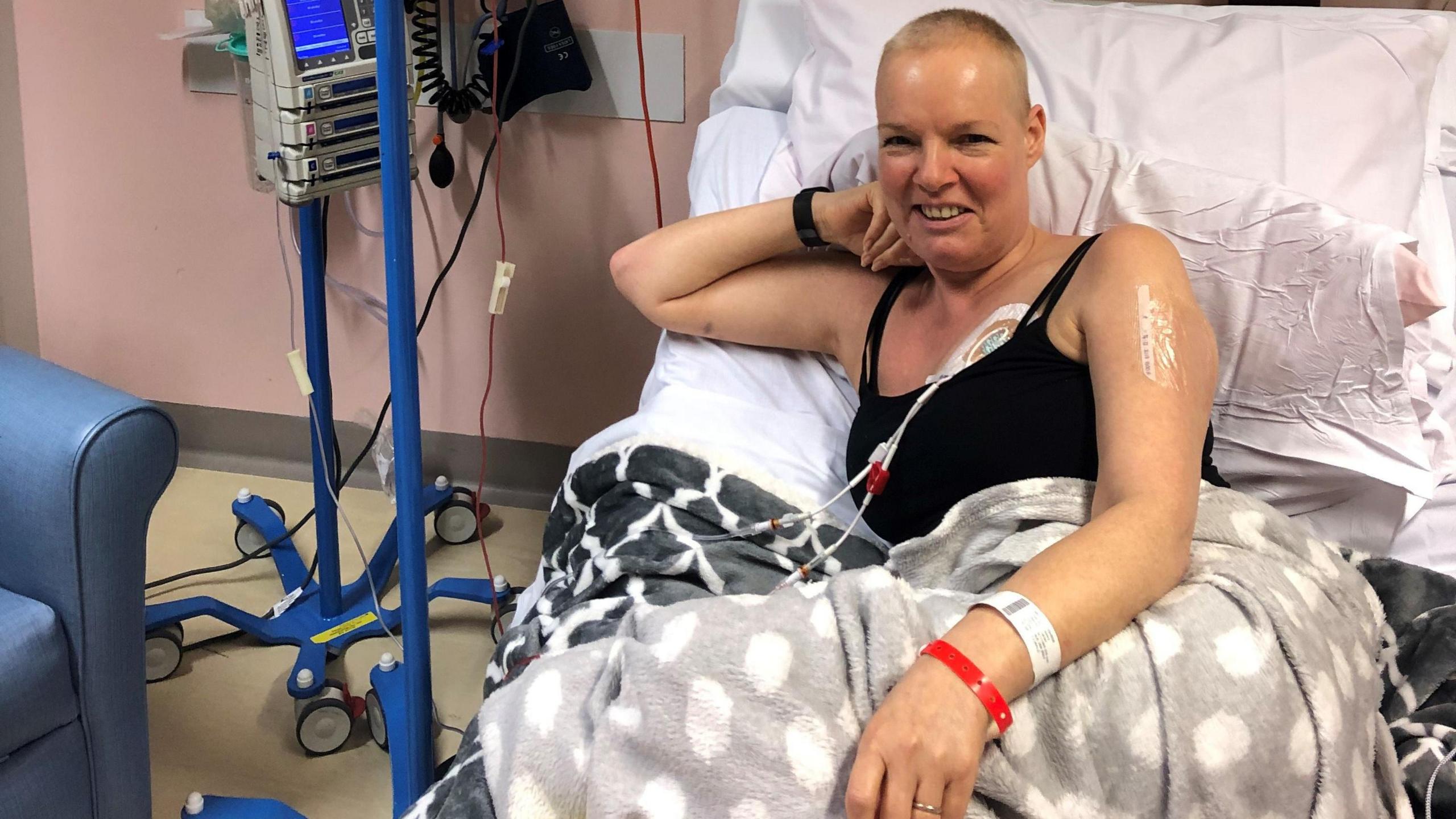 Lesley sitting up and smiling in a hospital bed, with a tube going into her chest. She has a bald head and is wearing a black vest and a blanket over her legs.