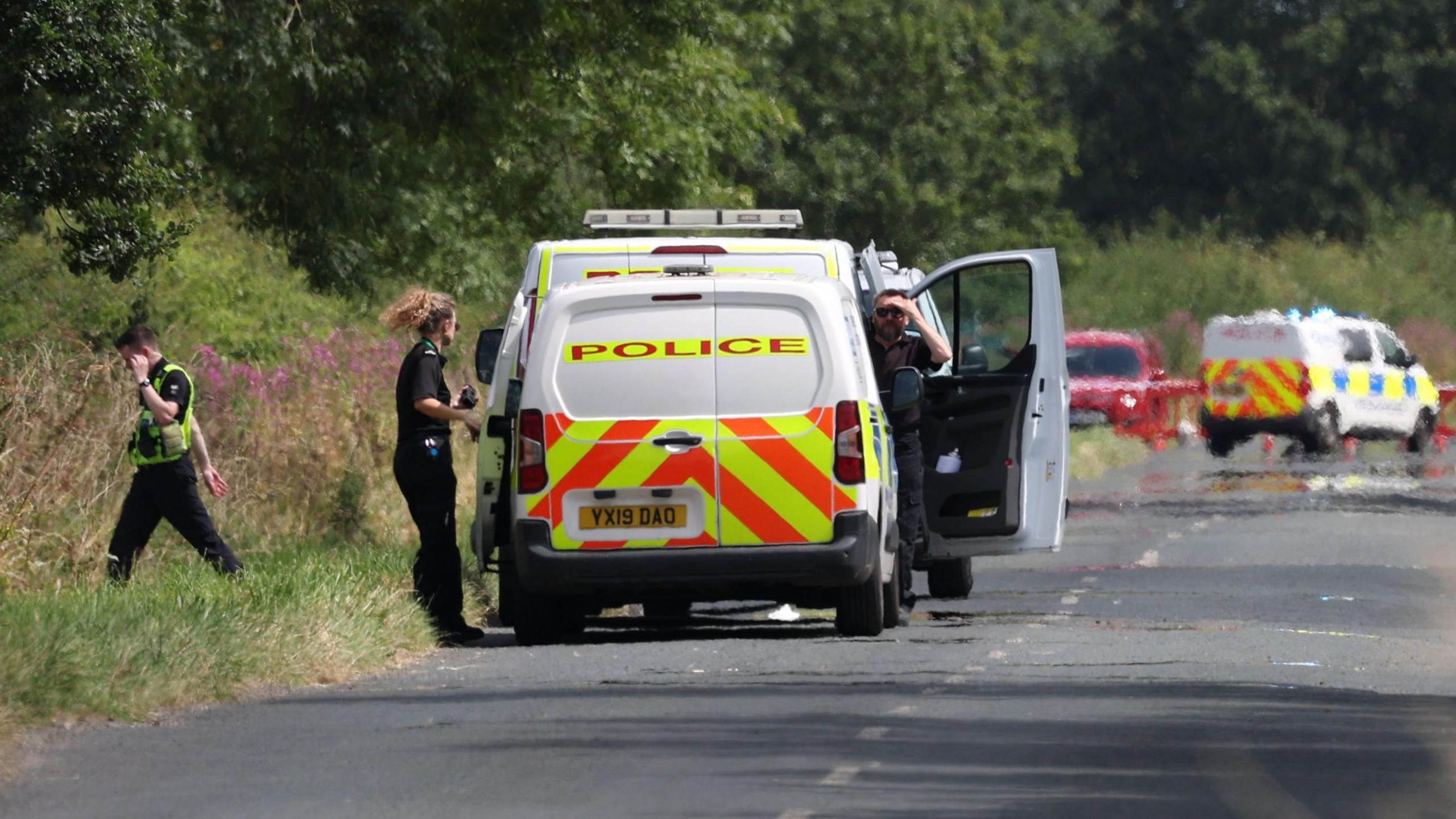 police van on a country road with heat haze on the road surface. A male officer wearing sunglasses shields his eyes and looks towards camera, a female officer stands on the near-side of the vehicle. A third officer wearing a hi vis vest is walking to the left of the van on a grassy verge.  