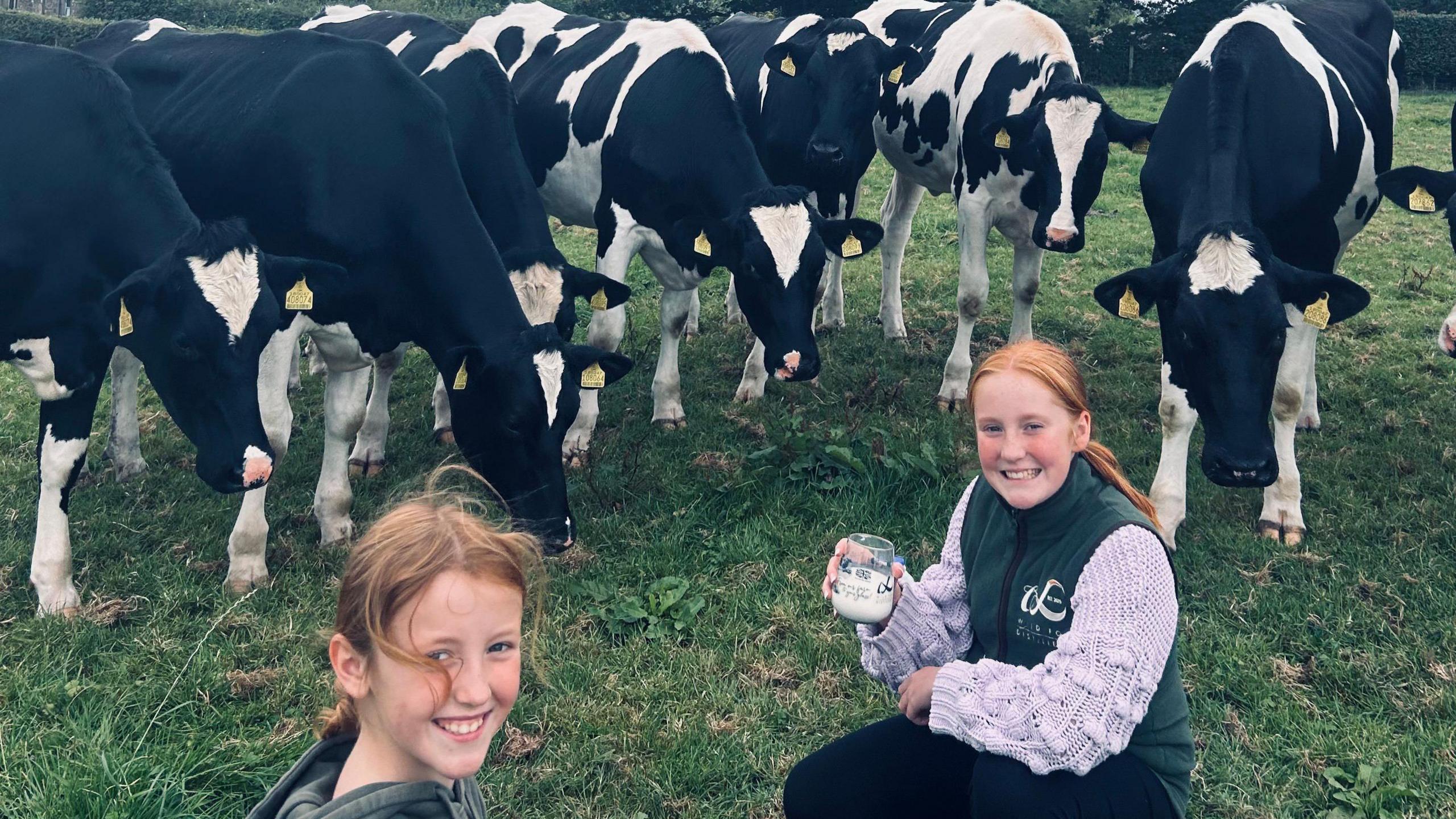 Annie and Betsy are standing with glasses of milk in front of the farm's herd of cows in a field. 