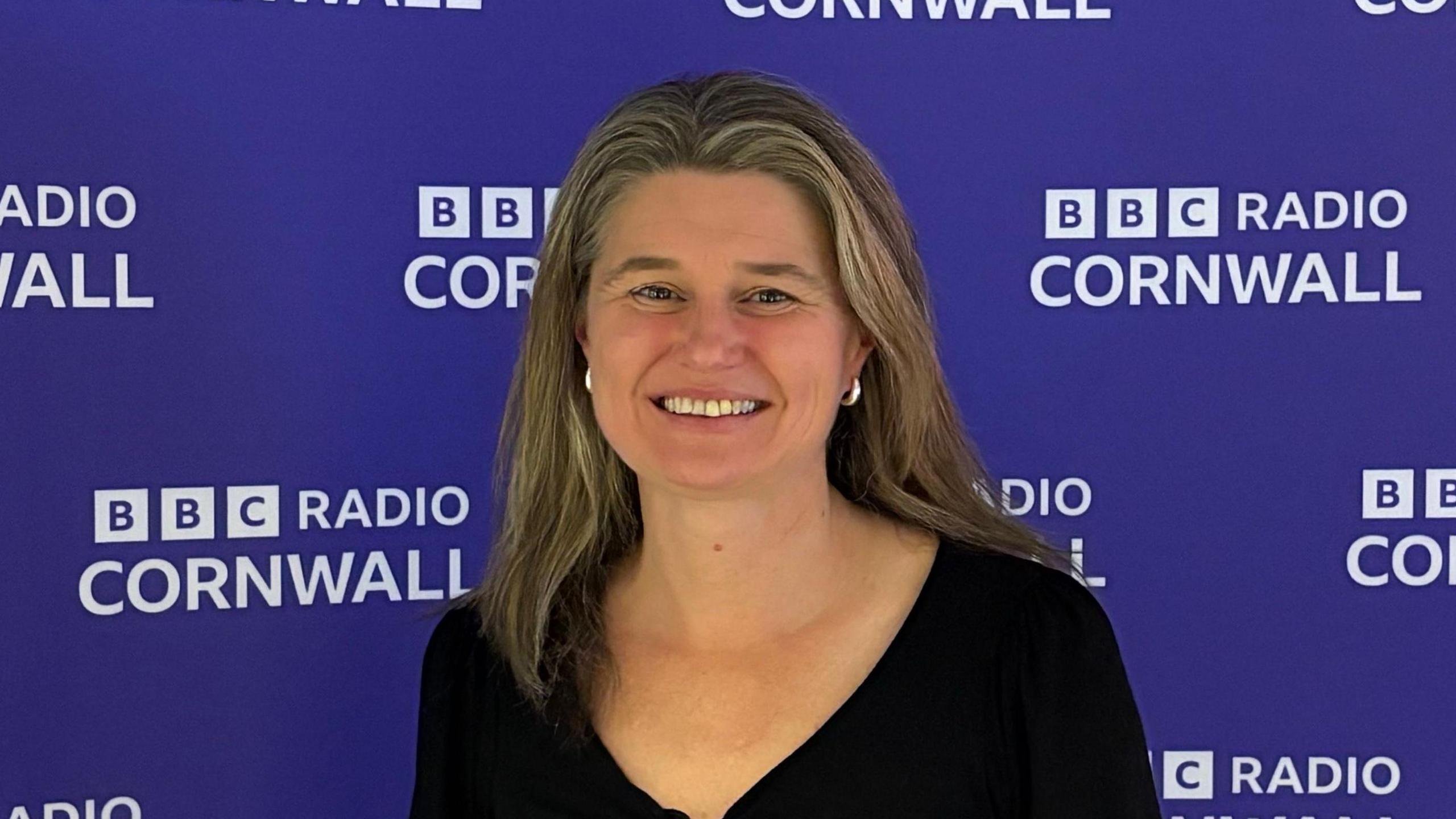 A man with long light brown hair wearing a black v neck top standing in front of a purple backdrop dotted with the BBC Radio Cornwall in white letters. 