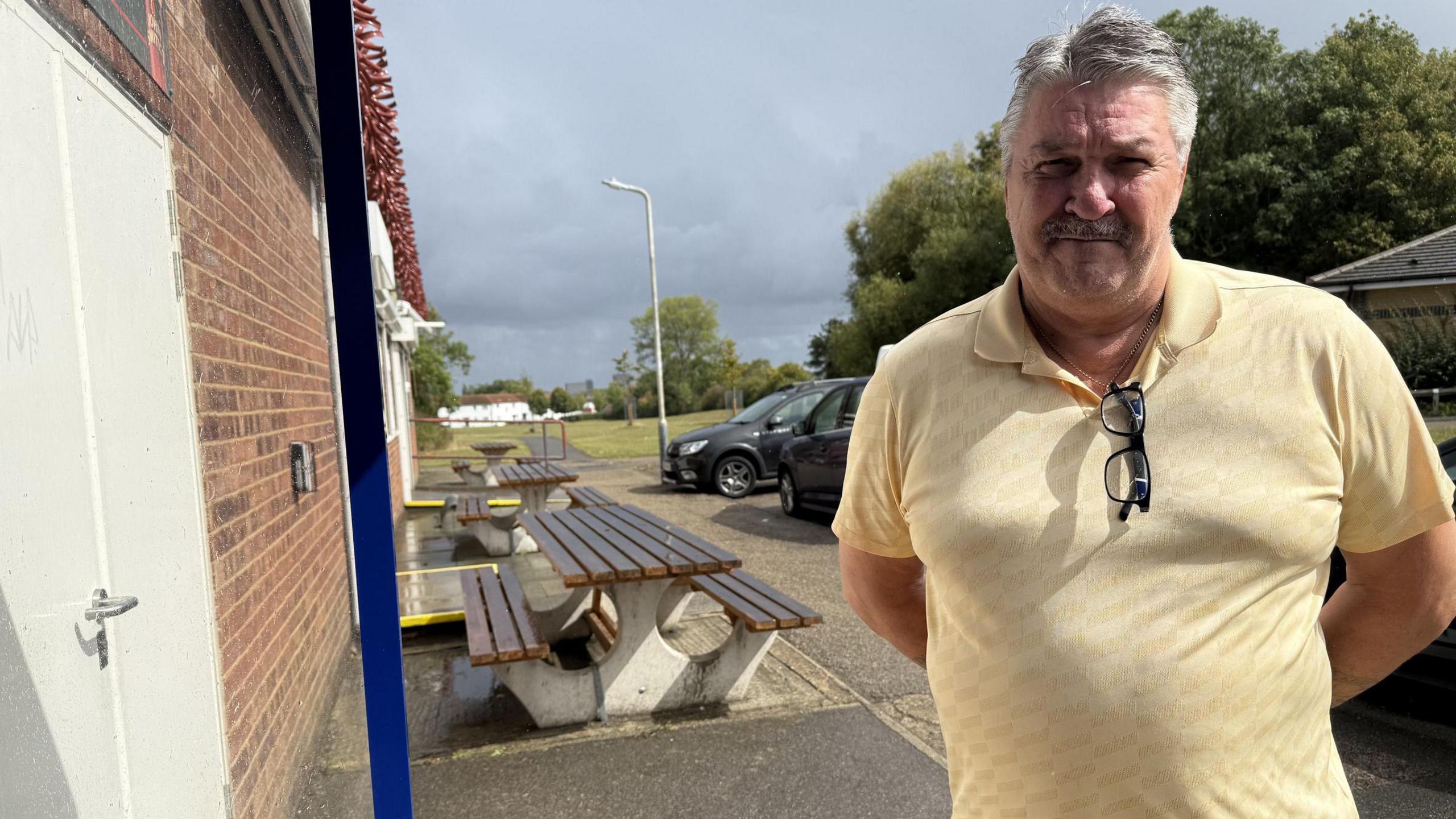 A grey-haired man with a moustache wearing a yellow polo shirt, stands with hands behind his back in front of some benches and cars