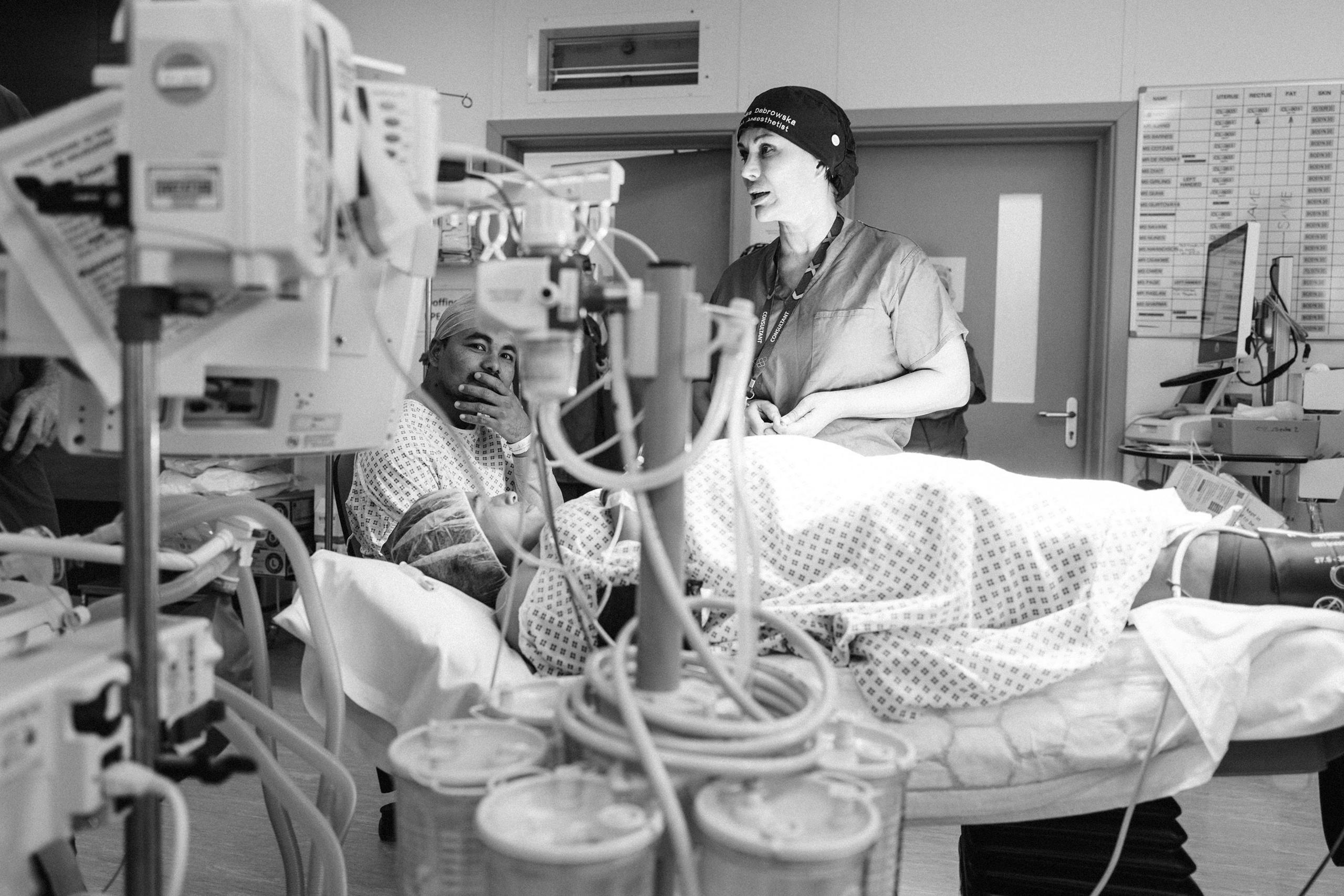 A patient appears to be lying down on a bed in a delivery room surrounded by medical equipment, with a healthcare professional standing nearby and her partner is sitting next to her with his hand covering his mouth.