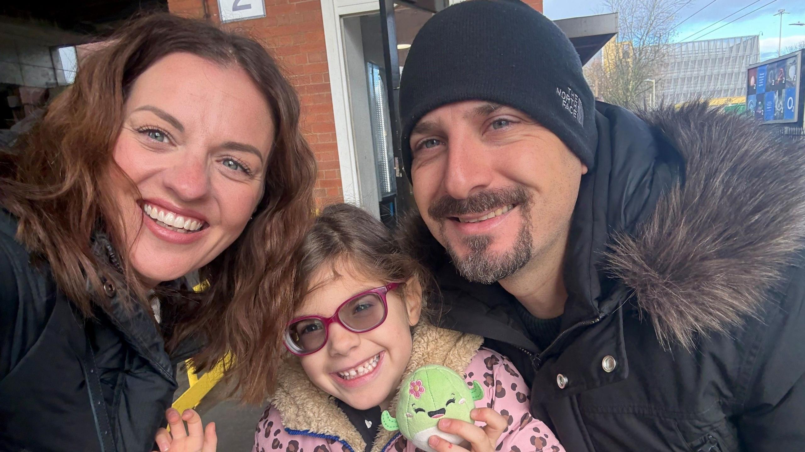 Three people all together smiling and looking at the camera. The young girl in the middle is holding a toy and has on a pink and brown leopard print coat, with purple glasses on. A woman is to the left, with long dark hair and a man to the right with a black hat on, and black coat with a furry hood.