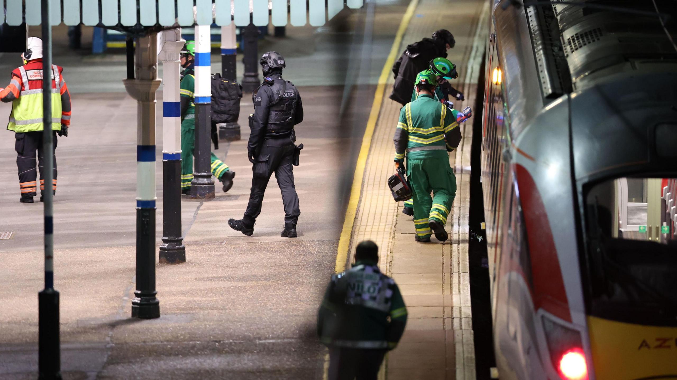 A train is parked at the station at night. People wearing various emergency kit uniforms mill around. Some all black, some green paramedics, and a few of them wear helmets.