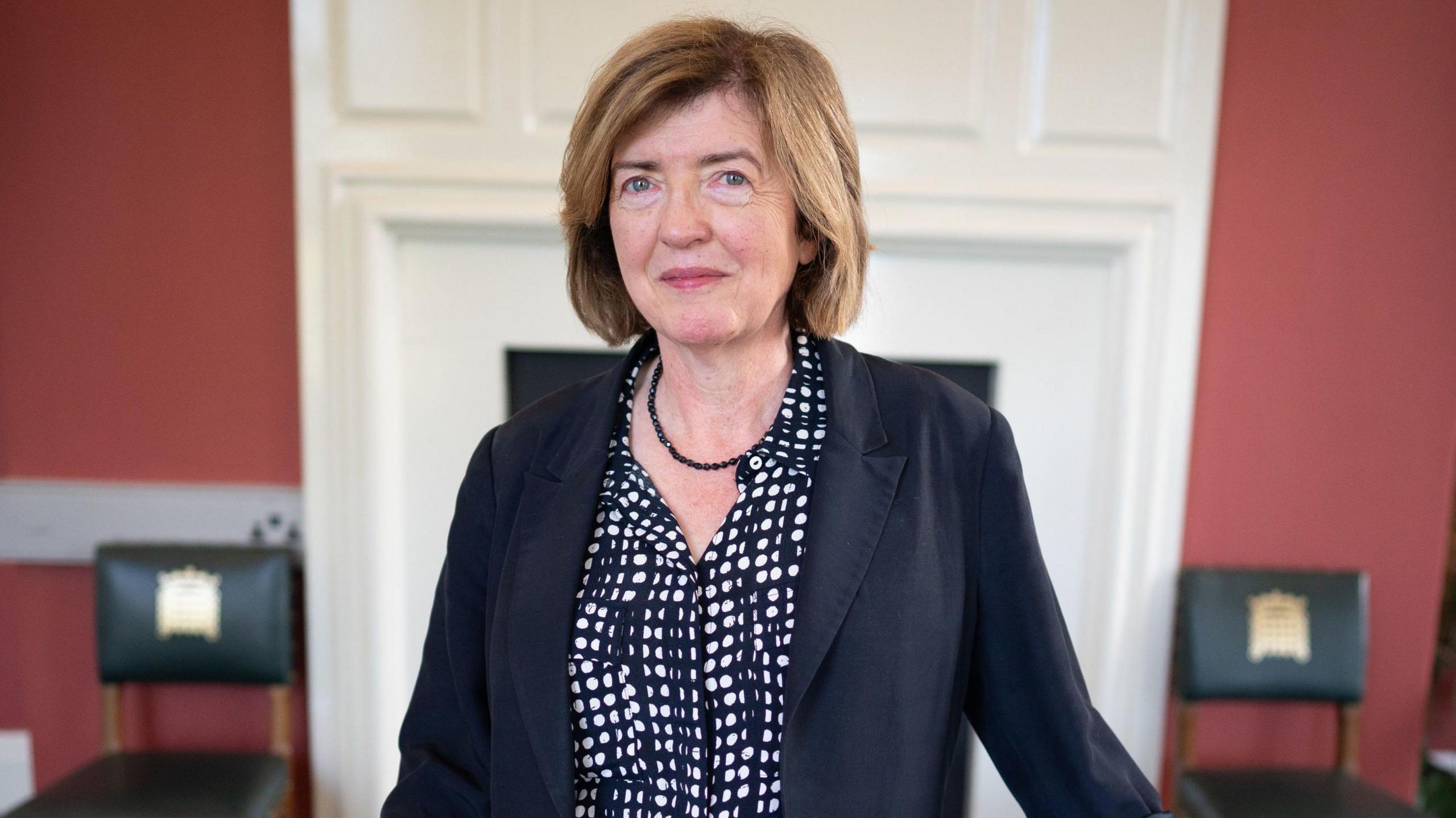 Sue Gray, wearing a dark blazer and dark shirt patterned with white circles, stands in a room with red and white walls, with two chairs in the background with the UK parliament logo on them