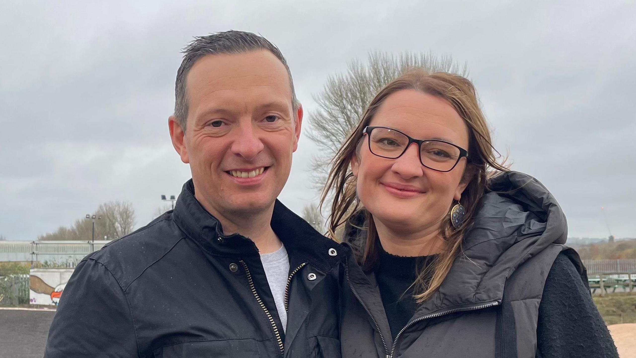 Jon and Laura at Exeter Eagles BMX Club. Jon is wearing a black jacket with a grey top underneath. He has short dark hair. Laura is wearing a black jumper with a black puffer coat over the top. Laura has brown hair and is wearing glasses.