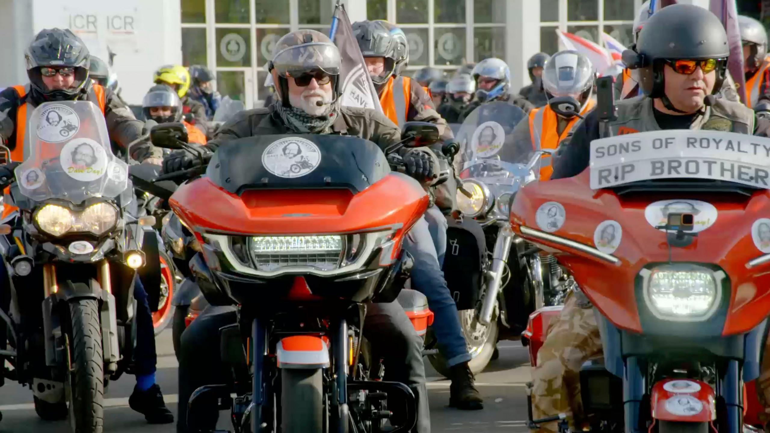 Si King is sitting on his motorcycle on Dave Day. He surrounded by other bikers. They are all wearing leathers and sunglasses. One of the signs on the handlebars of one of the biker's reads "Sons of Royalty, RIP brother".