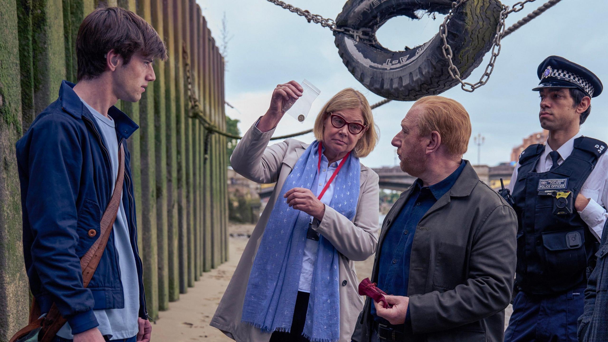 Jude, acting as Jake, stands on a beach with a group of detectives and police. Adrian Scarborough, acting as DI Max Arnold, holds a red velvet bag in his hand and looks at a small bag with a coin being held by his female colleague. She wears a long blue scarf, beige overcoat and red glasses. A man in police uniform looks on. There is a busted tyre hanging overhead.
